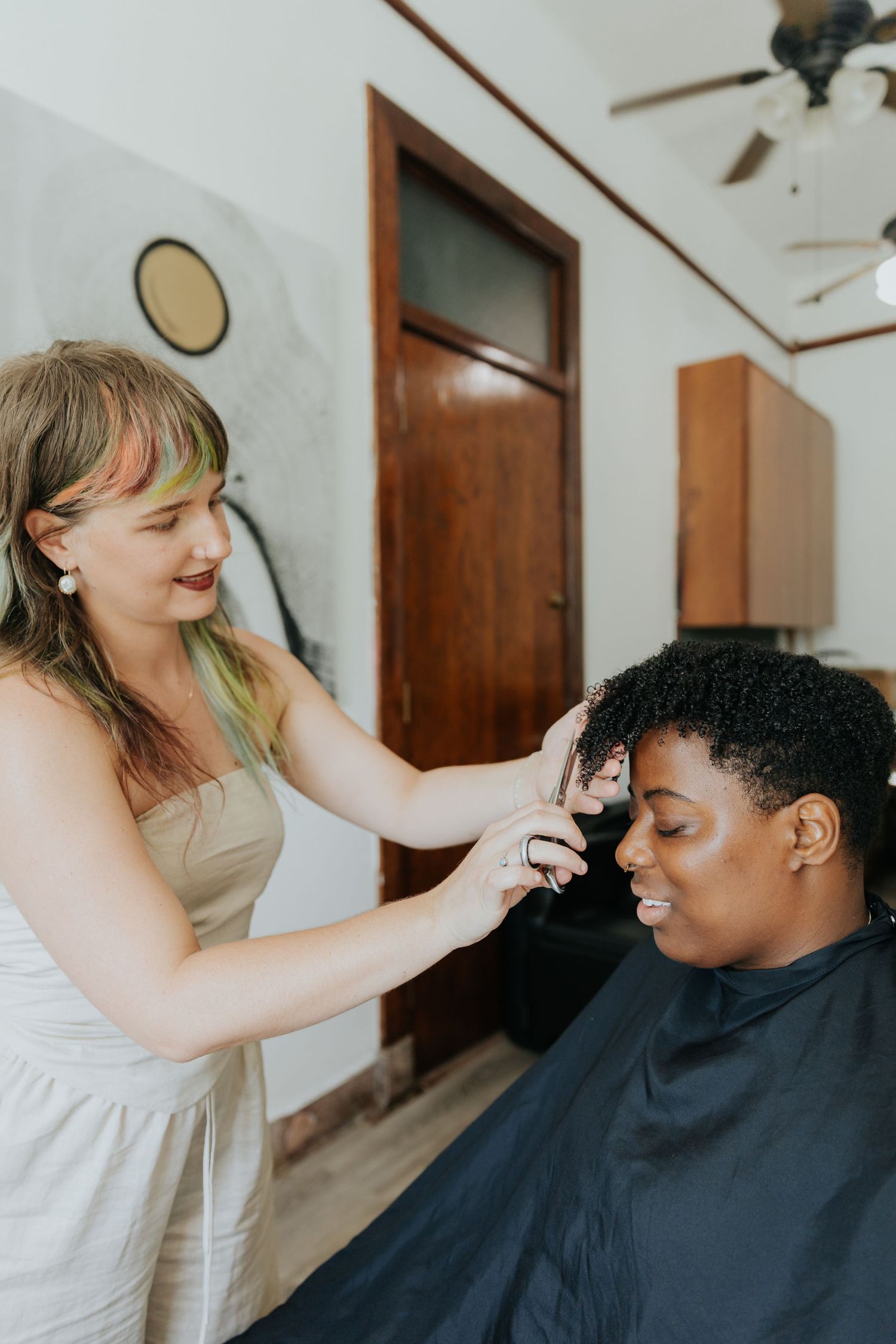 A hairstylist works on styling a client's hair while wearing a black cape in a bright indoor setting.