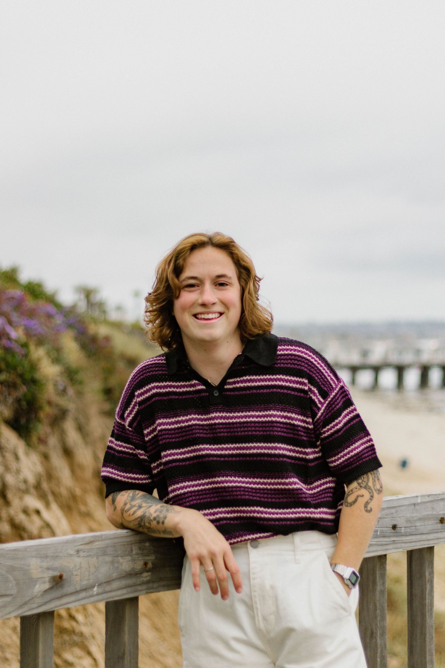 Person in striped sweater poses casually along wooden beach boardwalk railing on cloudy day.