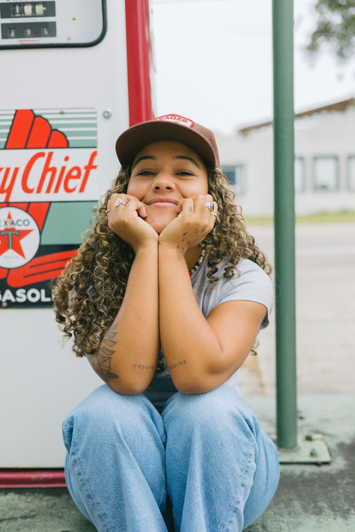 A person in a baseball cap and jeans sits casually on a curb in front of a convenience store.