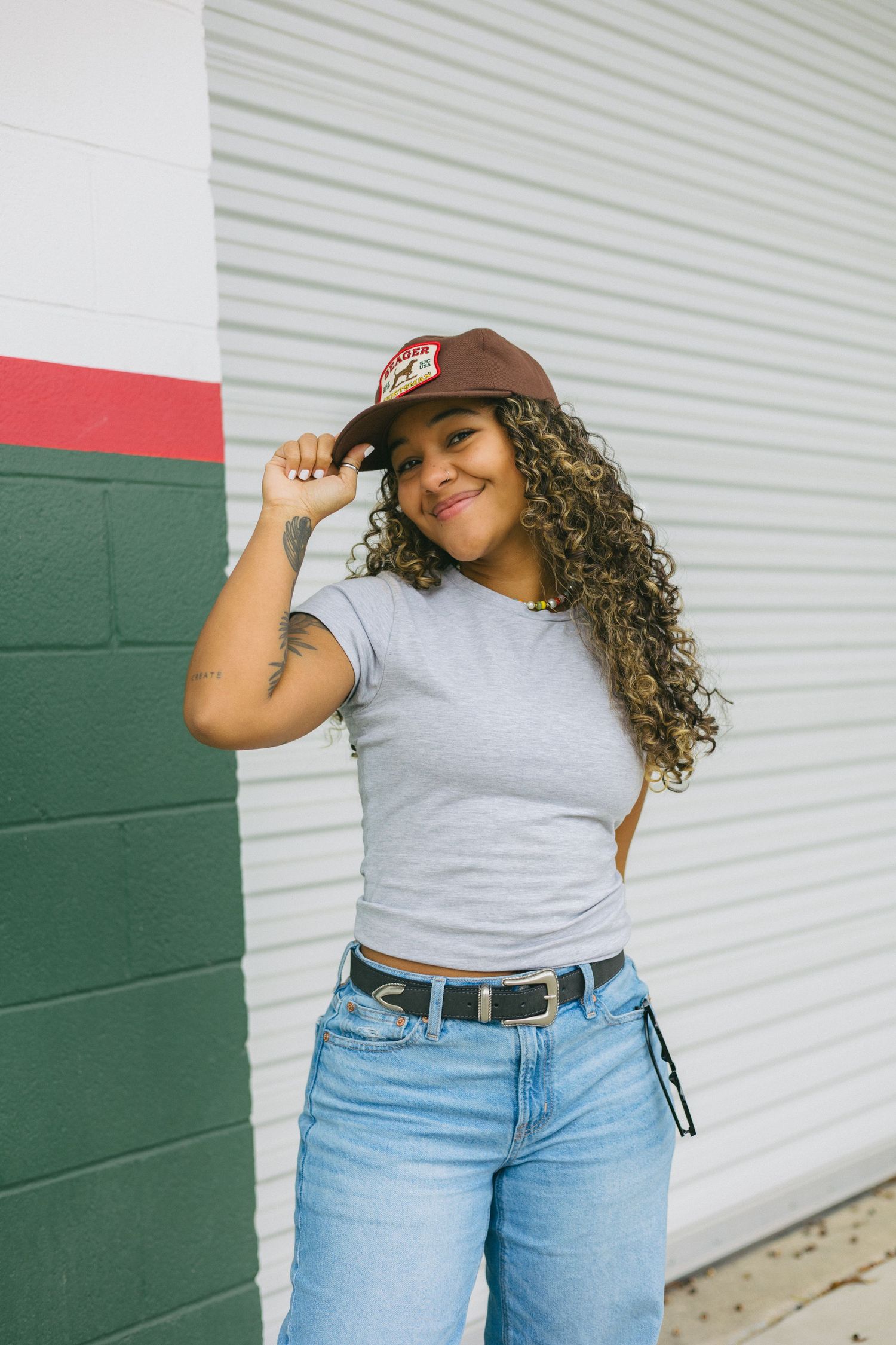 A person in a gray t-shirt and light blue jeans poses casually against a white and green wall background.