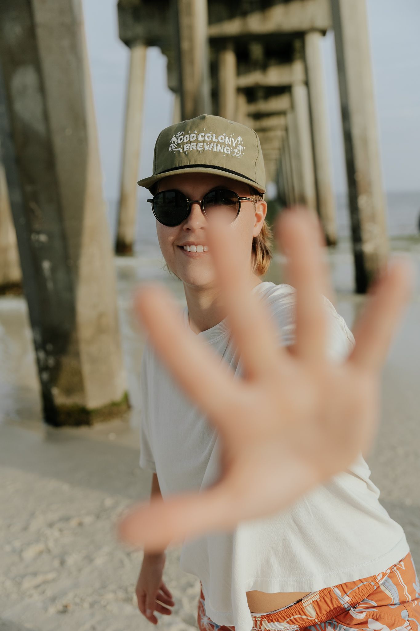 Person in a baseball cap and sunglasses gestures with their hand on a sunny beach near a pier.
