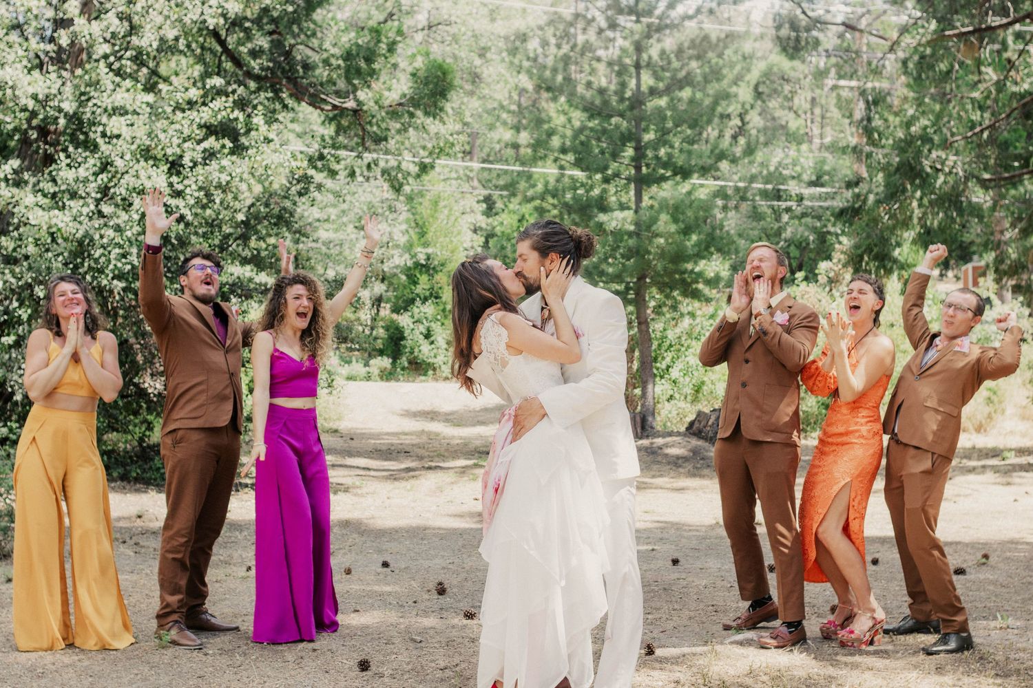 Wedding party poses for photos in coordinated brown and orange attire under green trees.
