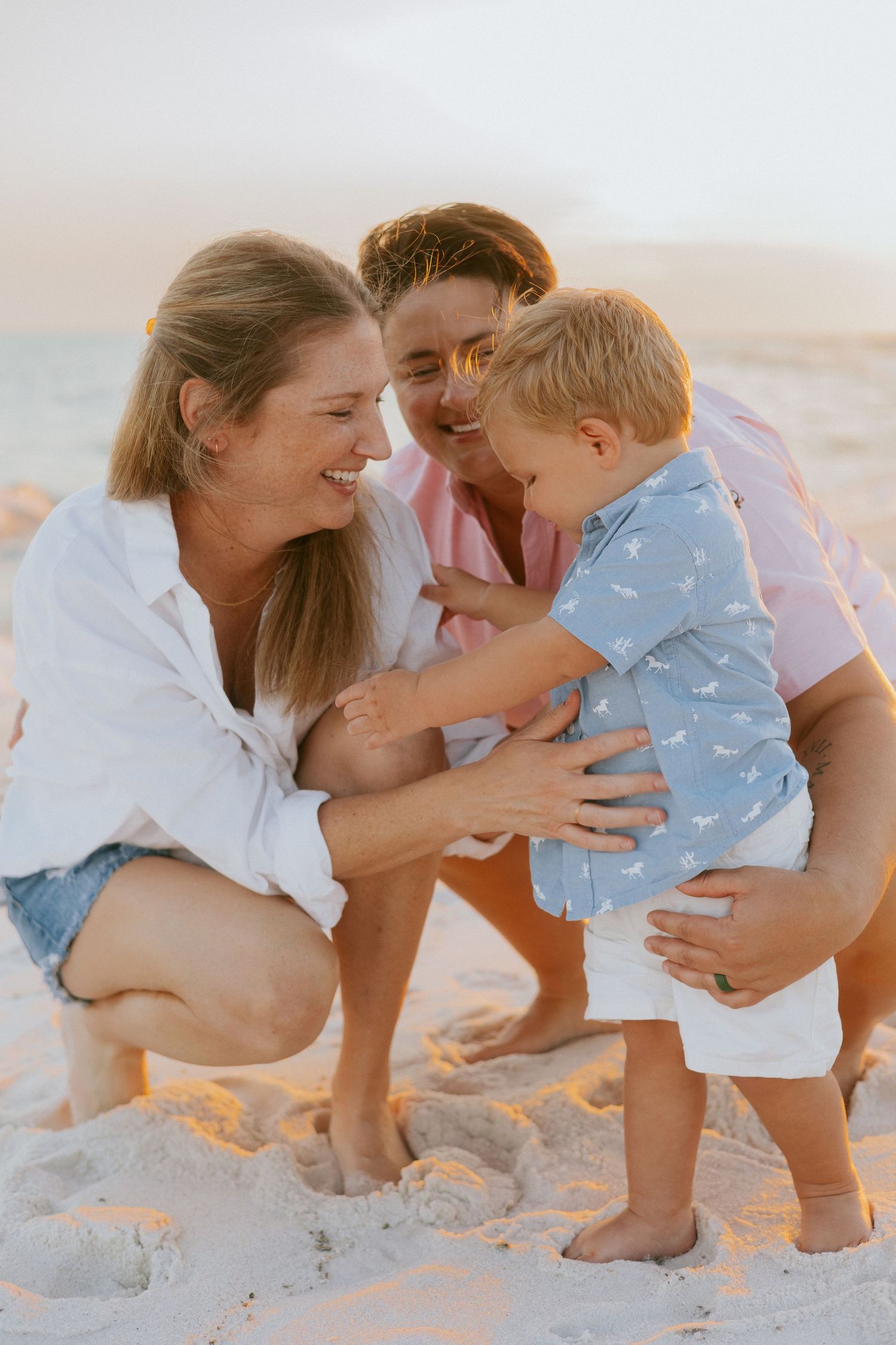 Two women and a toddler boy enjoy a joyful family moment together on a sandy beach at golden hour.