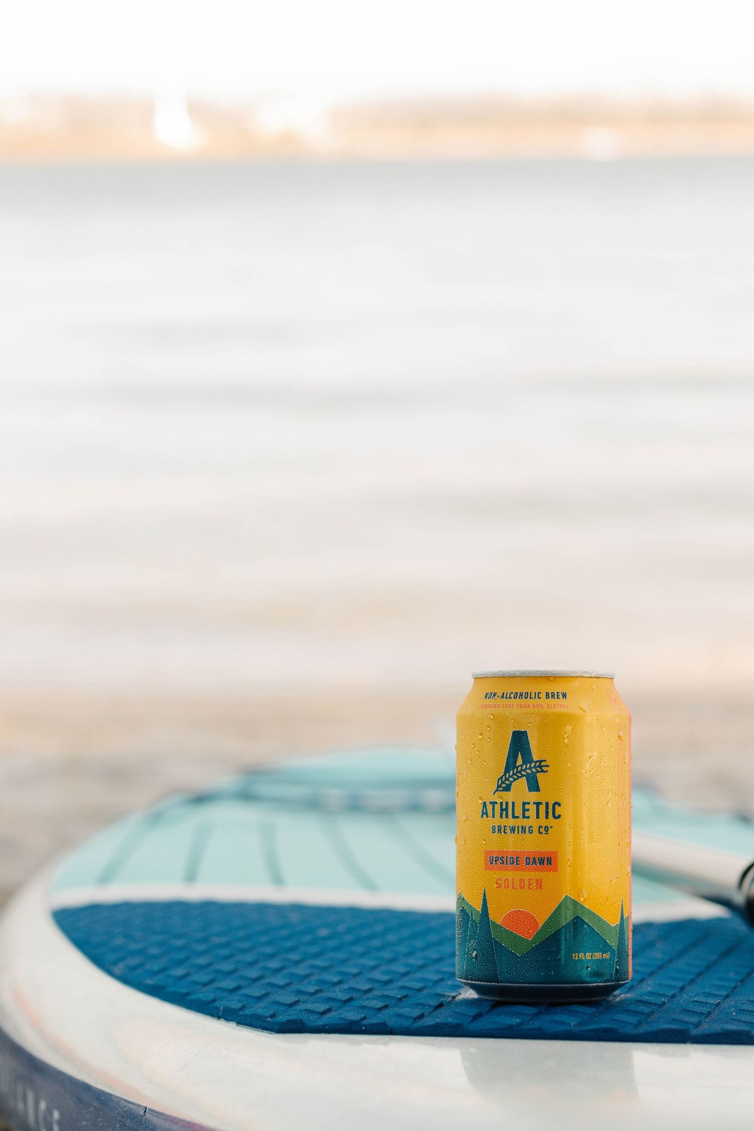 Yellow Athletic Brewing beer can sits on a blue surface with blurred background at sunset.