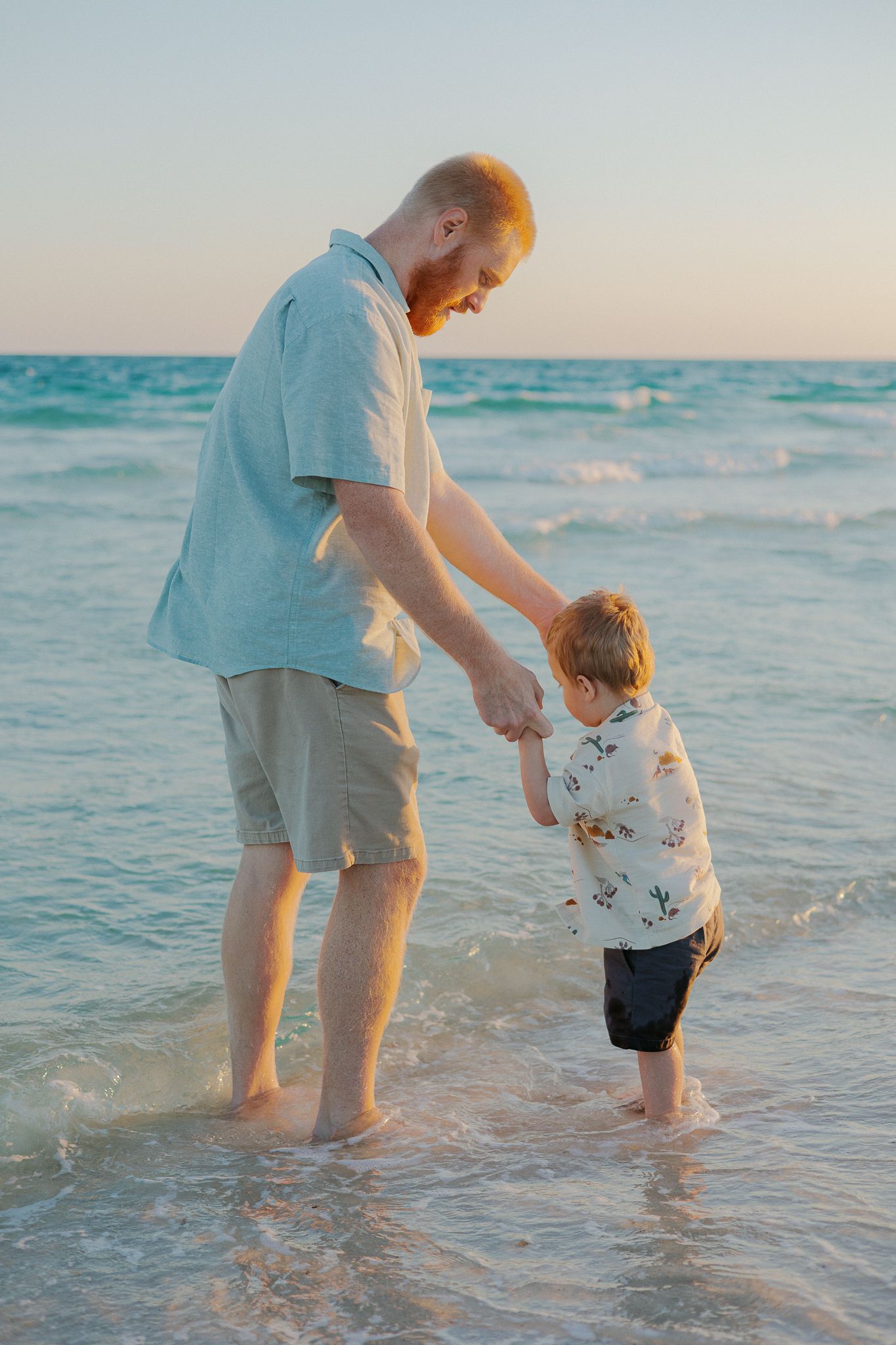 A father holds his toddler's hand while wading in ocean waves at sunset on a sandy beach.