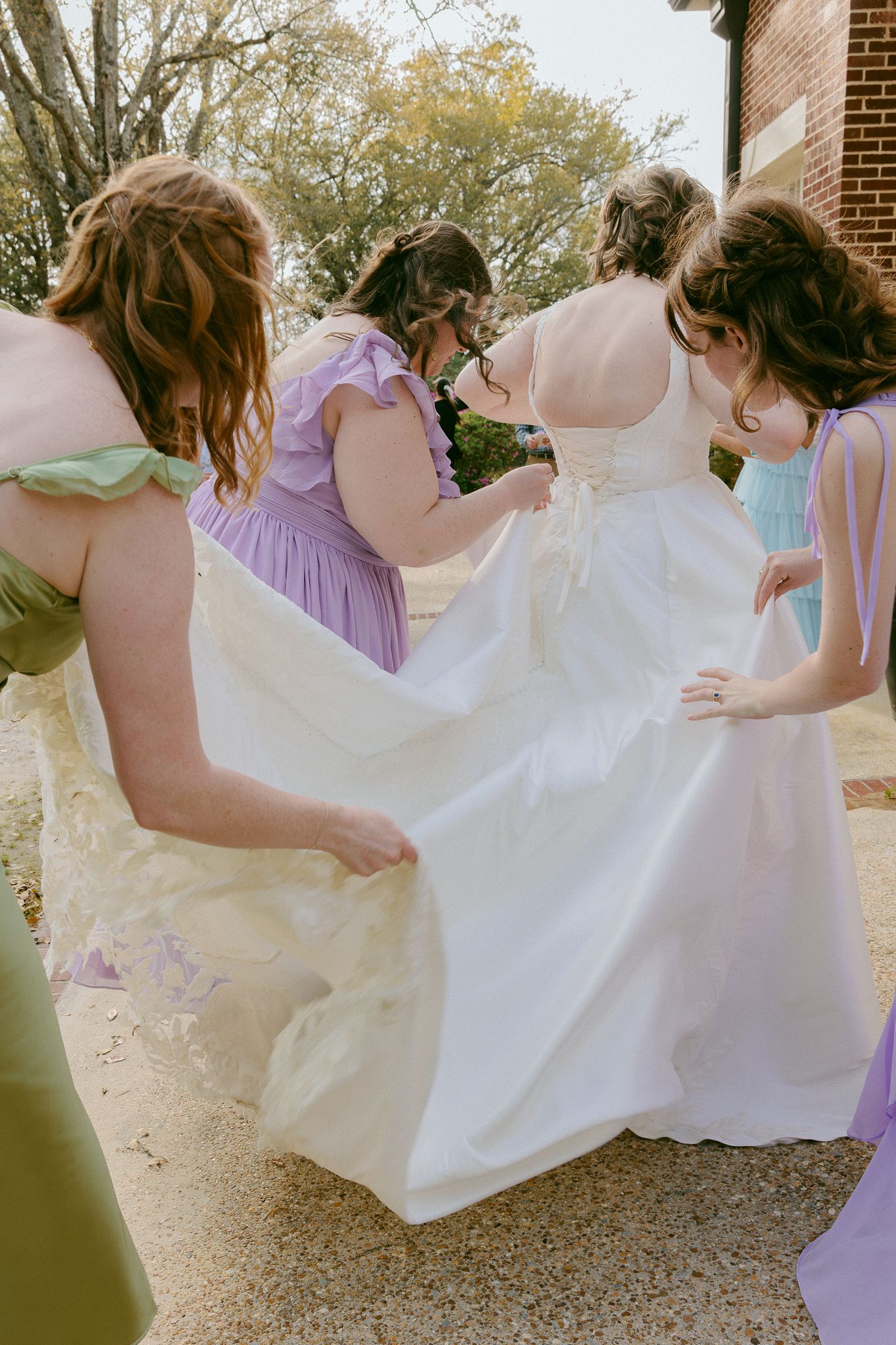 Bridesmaids in colorful pastel dresses help adjust the bride's white wedding gown train outdoors.