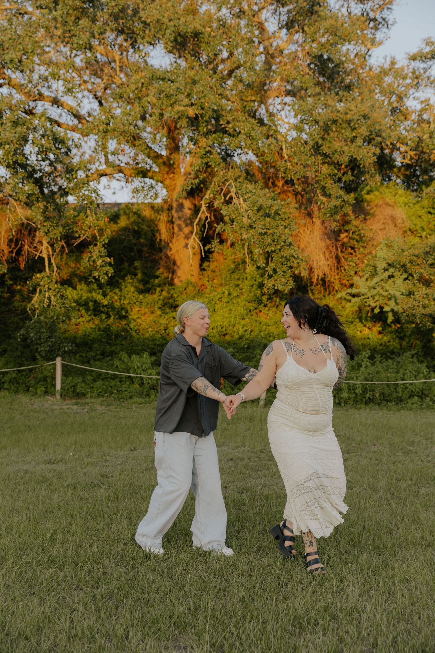 A couple dances together in a grassy field at sunset with trees in the background.