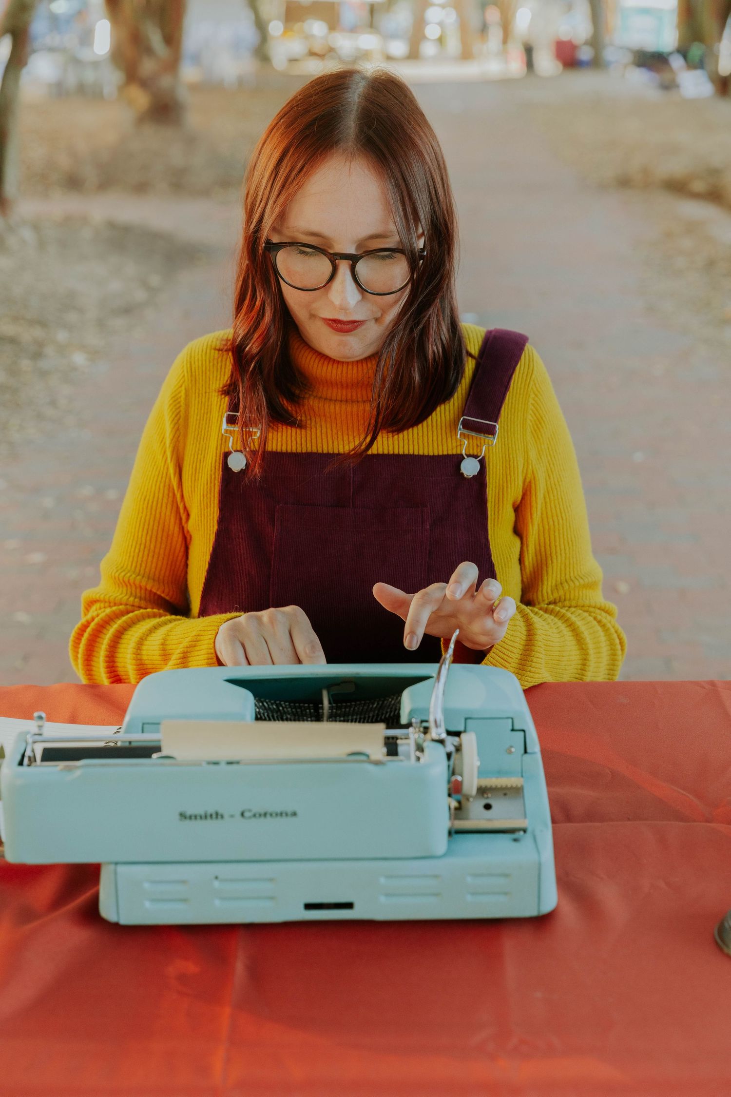 Writer in yellow sweater and brown overalls types on vintage blue Smith Corona typewriter at red table outdoors.