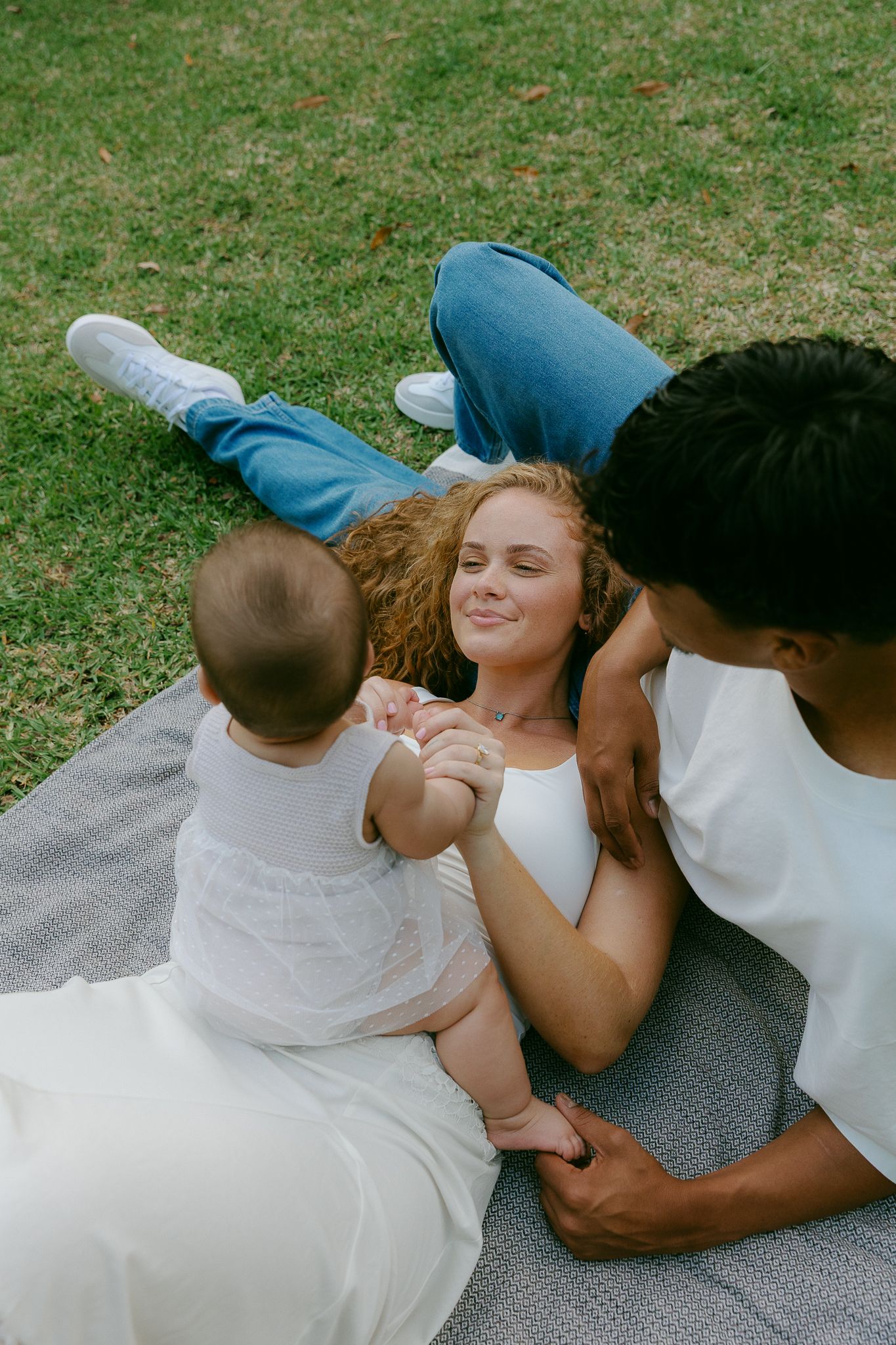Young family relaxing on a blanket in the park, mother smiling at baby while father sits nearby on green grass.