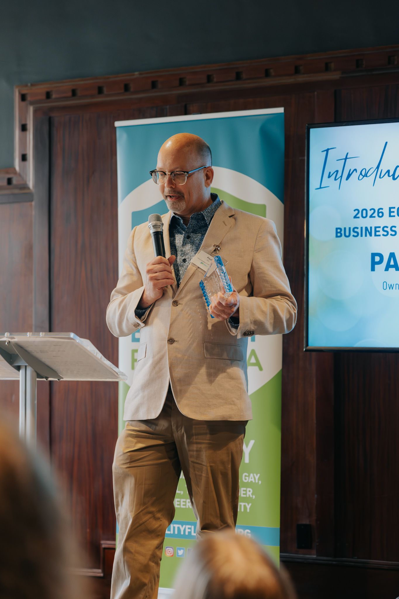 A speaker in a tan suit presents on stage at a business conference with digital screens visible in the background.