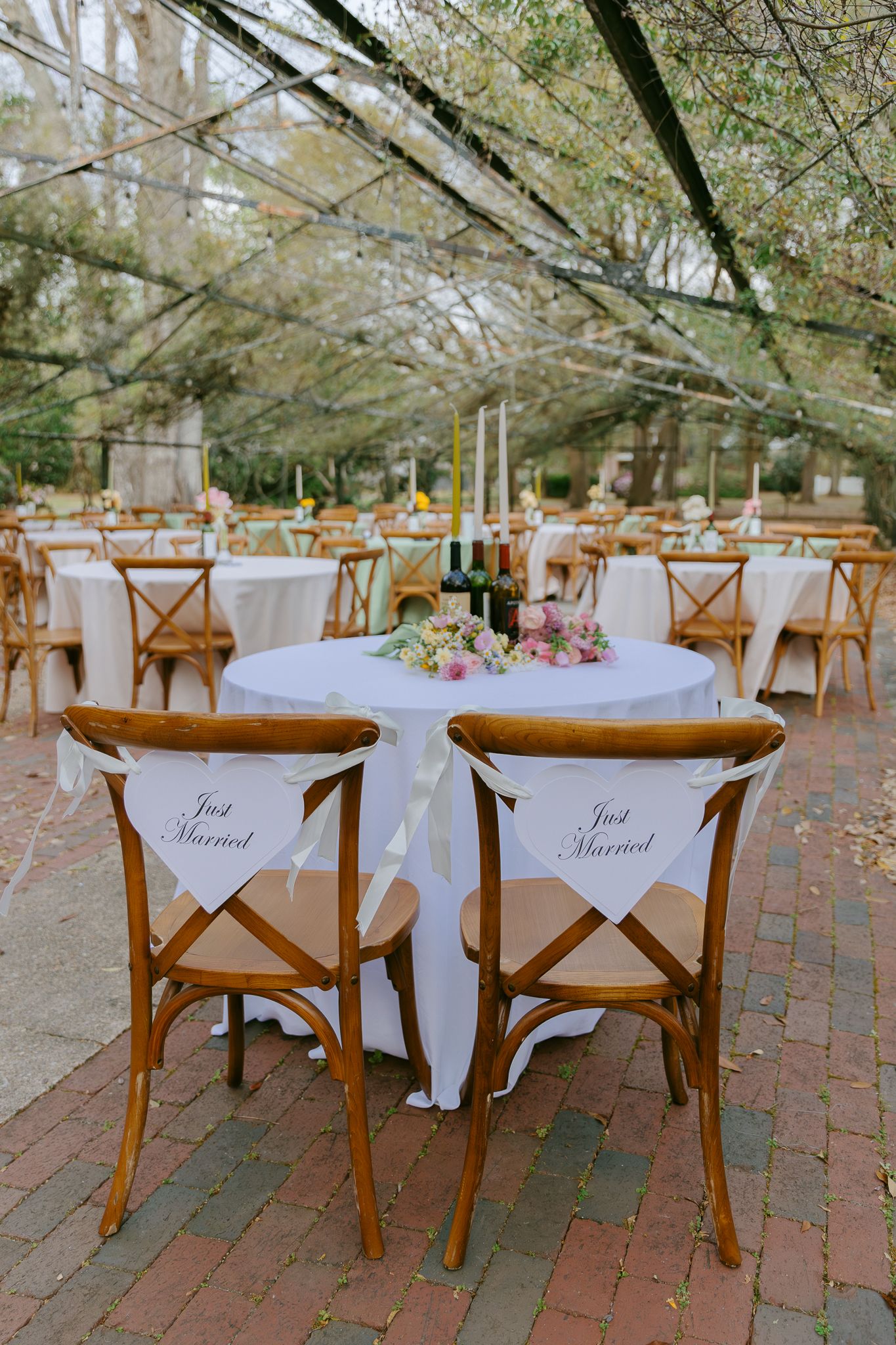 Outdoor wedding reception with Just Married signs on wooden cross-back chairs at a white-draped sweetheart table.