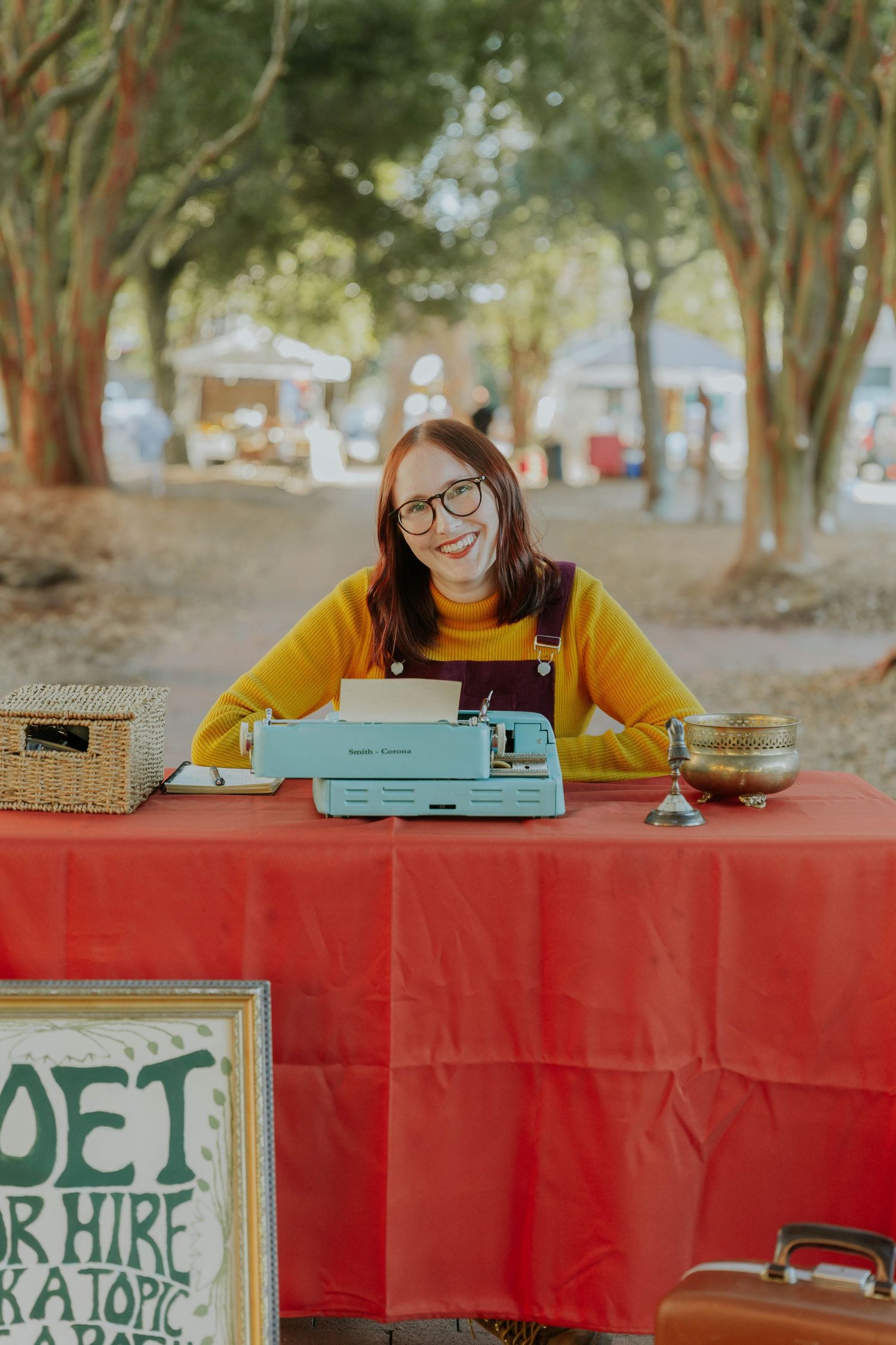 A person in a yellow sweater sits at a red-covered table outdoors with books and a vintage typewriter.