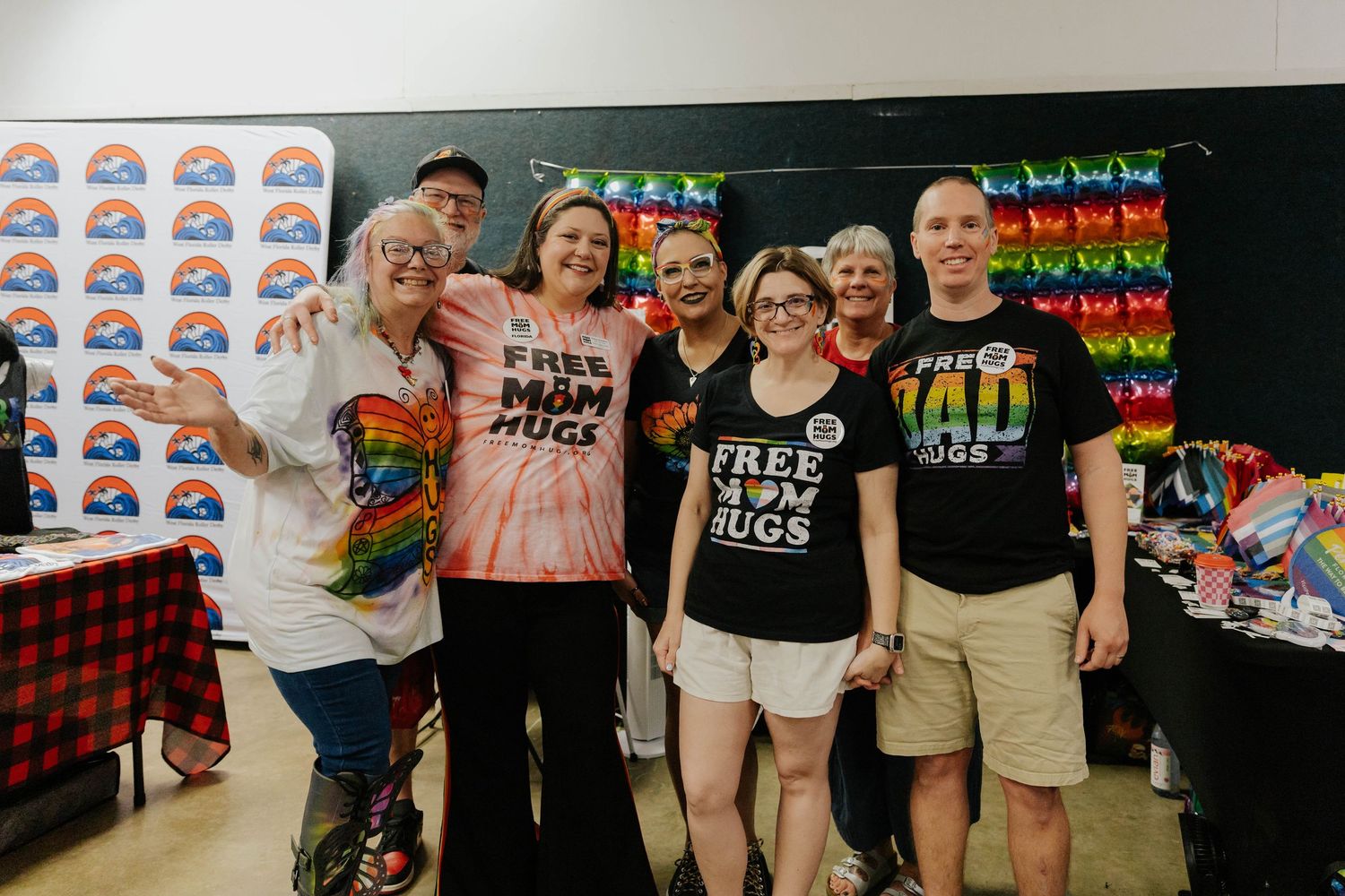 A group of people pose together at a Pride event with rainbow decorations and merchandise visible in the background.