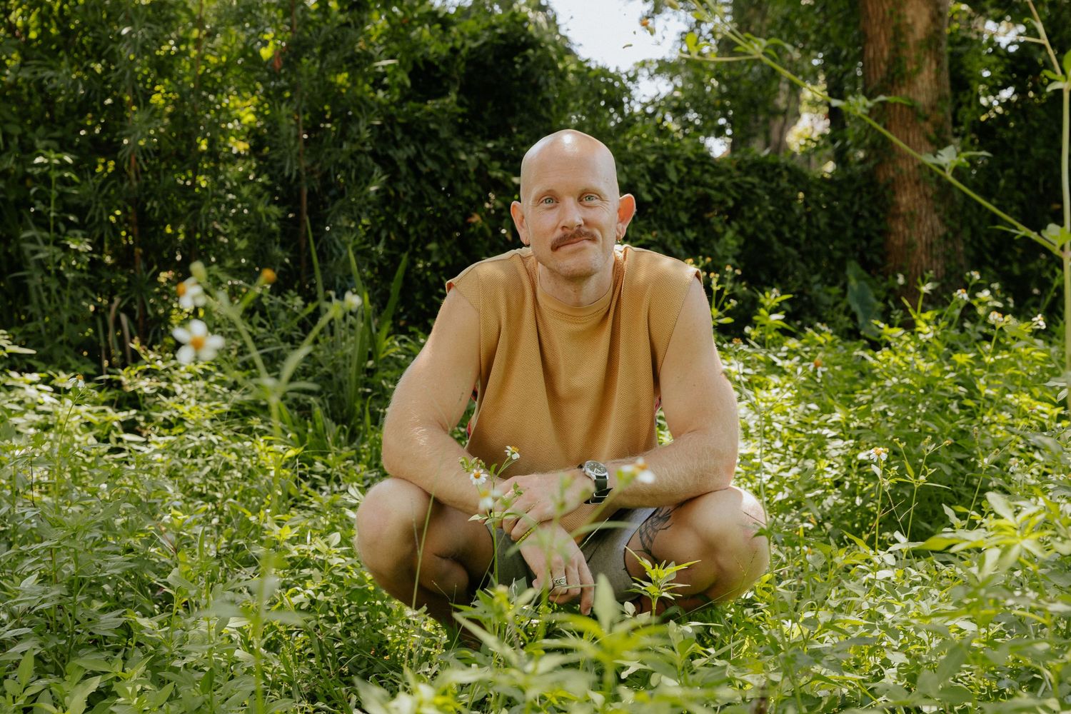 Person in a tan sweatshirt crouches in lush green garden setting on a sunny day.