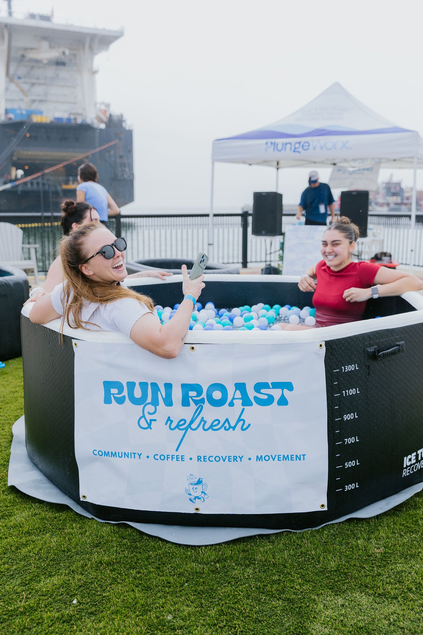 People relax in an ice bath setup with a 'Run Roast & Refresh' banner at an outdoor event near a ship.