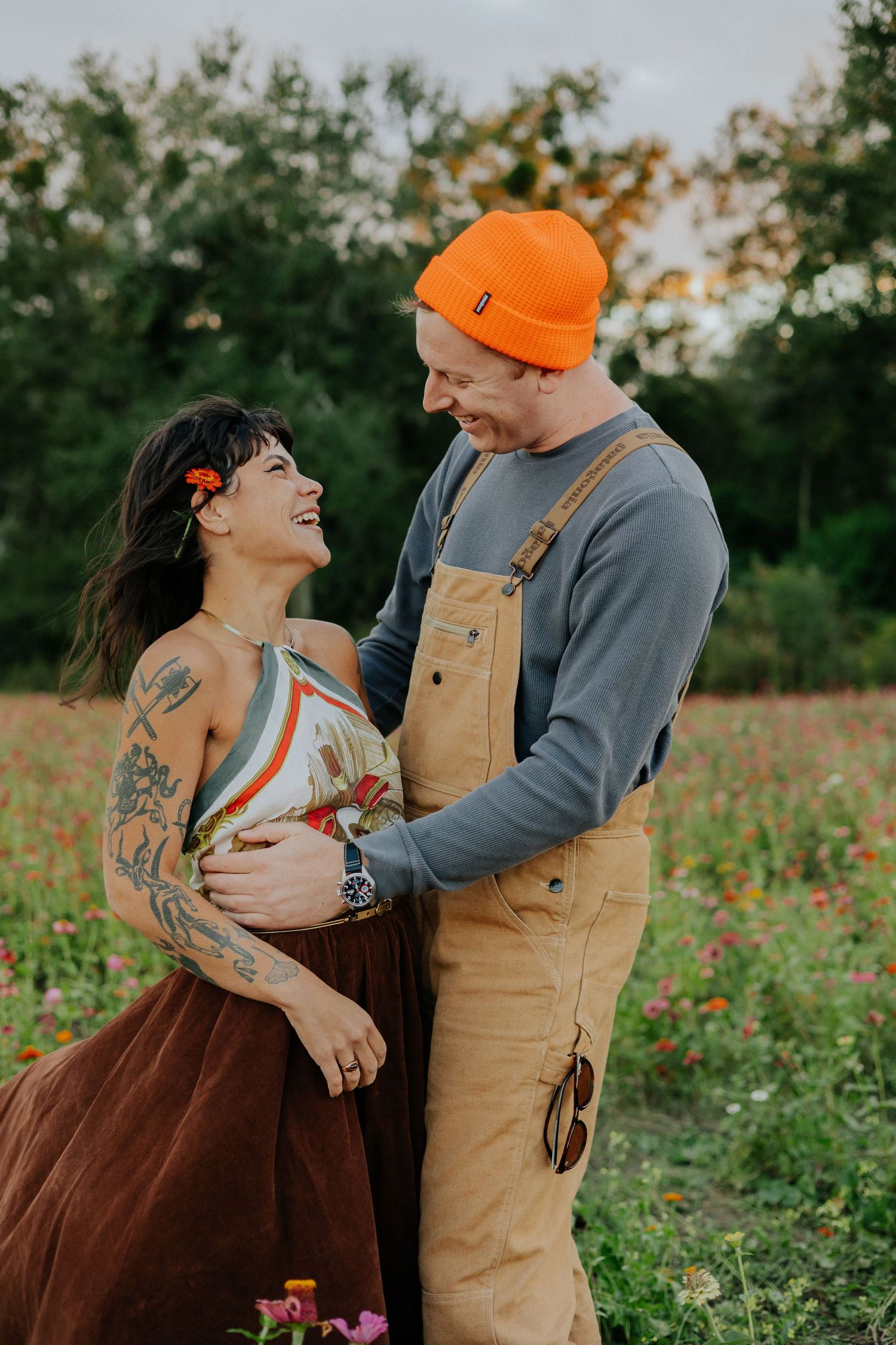 A couple shares an intimate moment in a wildflower field, with one wearing tan overalls and an orange beanie.