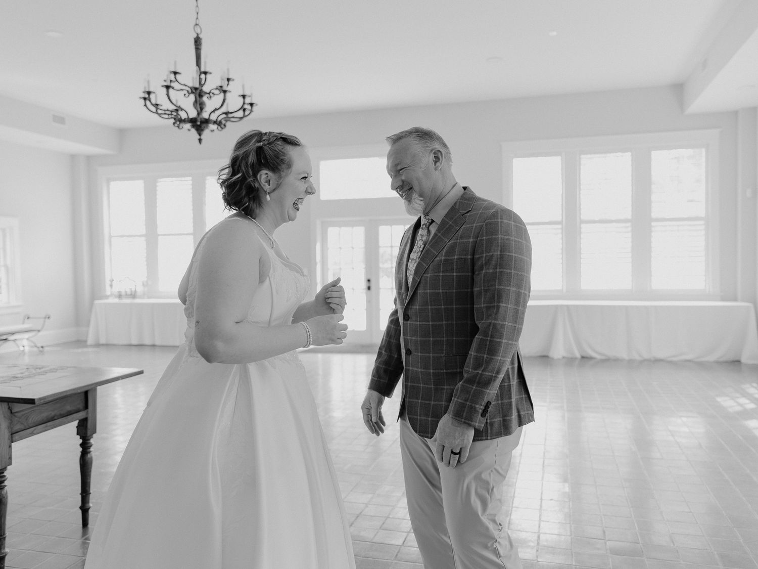 Black and white photo of a bride in white gown laughing with a man in plaid blazer inside an elegant venue.
