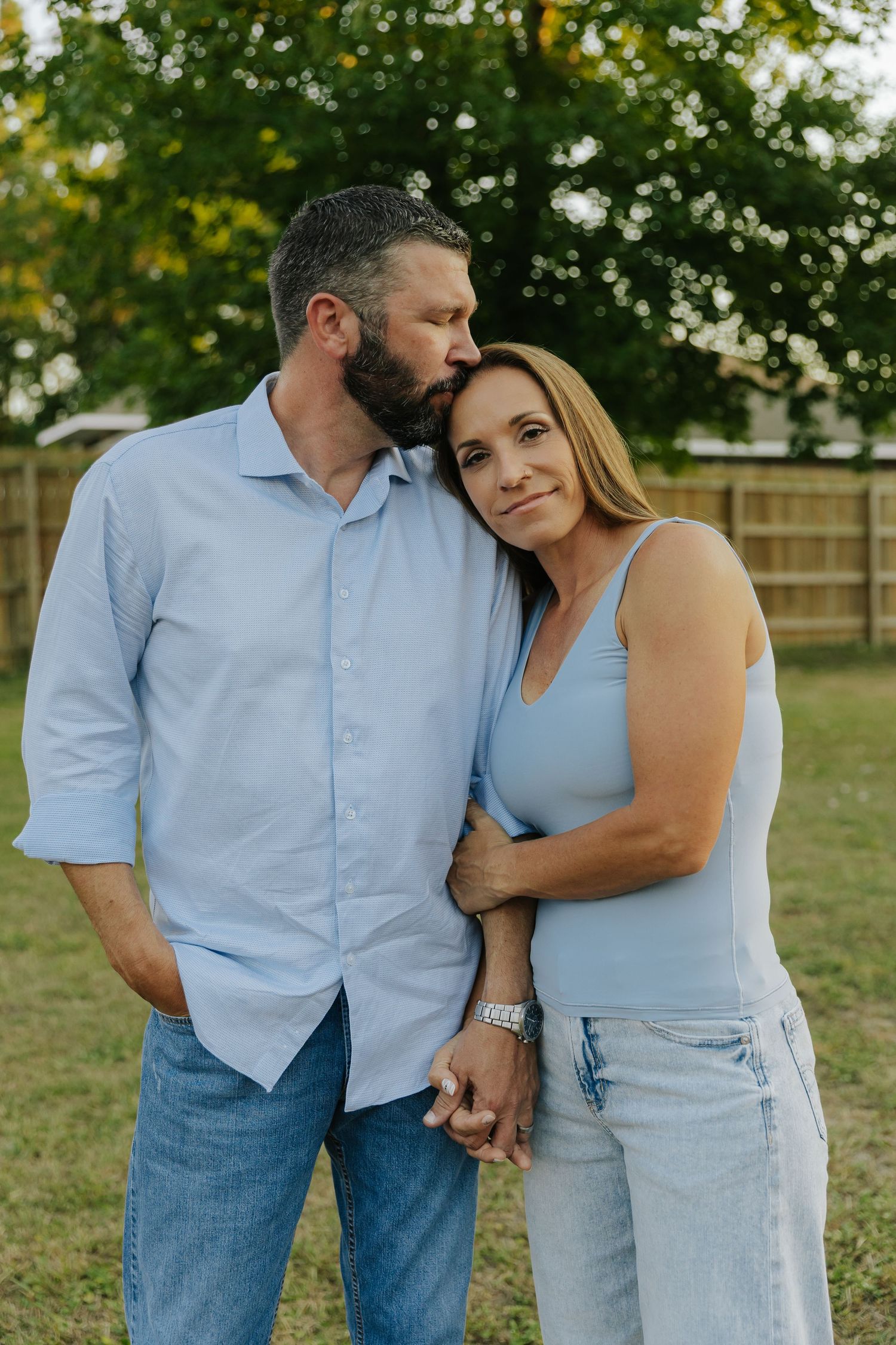 A couple wearing light blue and denim share tender moments in a grassy outdoor setting during a photo session.