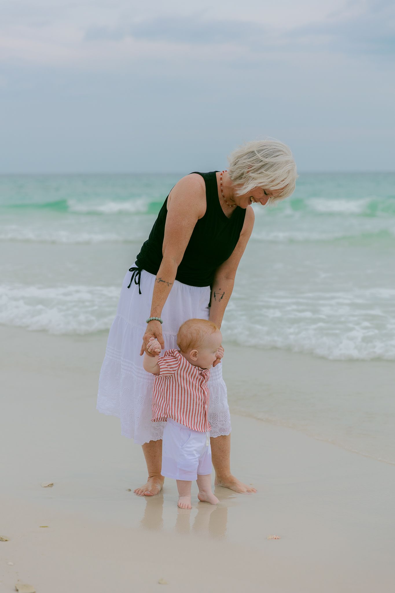 Grandmother helping baby take first steps on a sandy beach with turquoise ocean waves in the background.