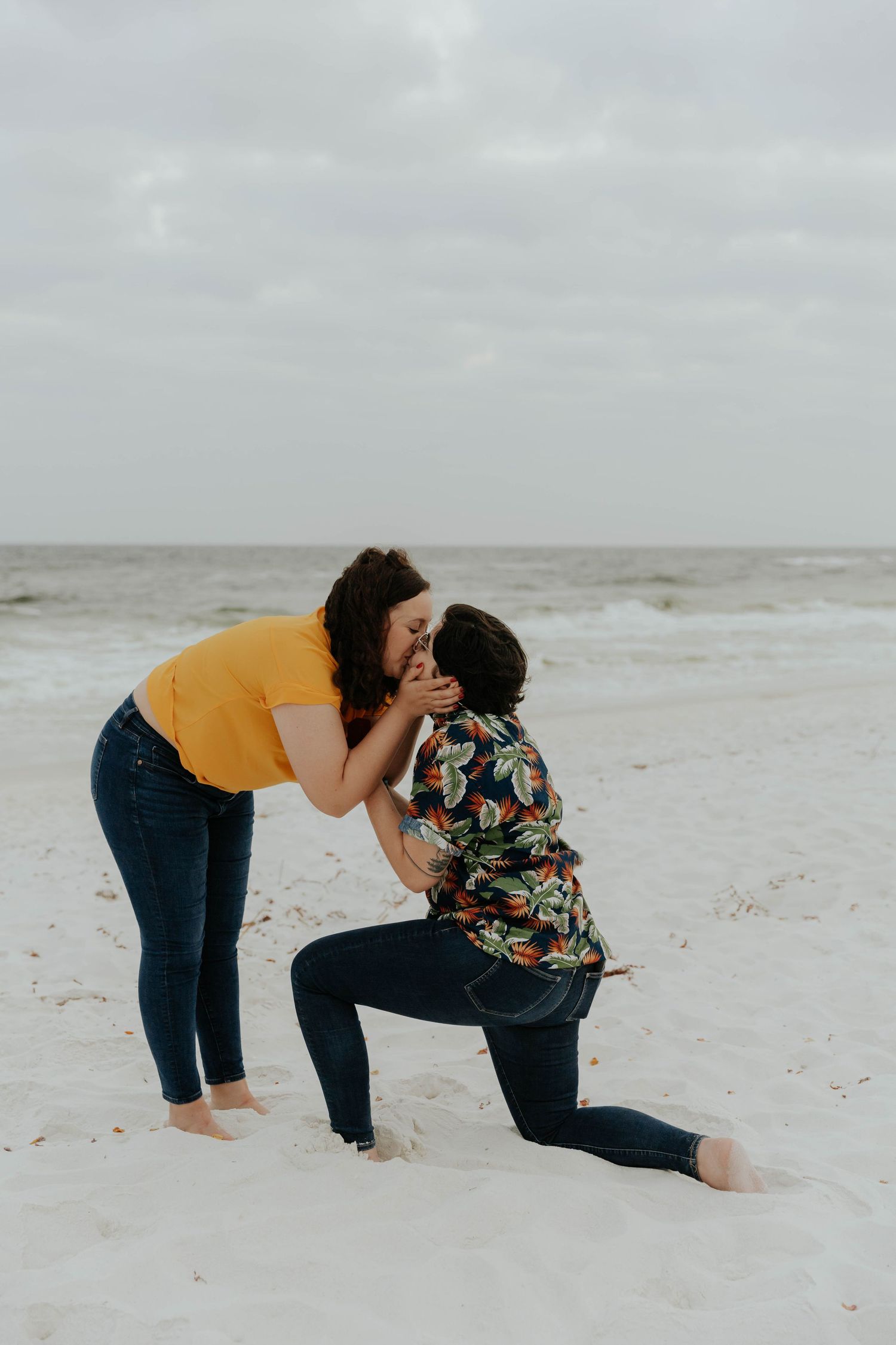 A couple shares an intimate moment on a cloudy beach as one kneels in the sand while wearing a floral shirt.