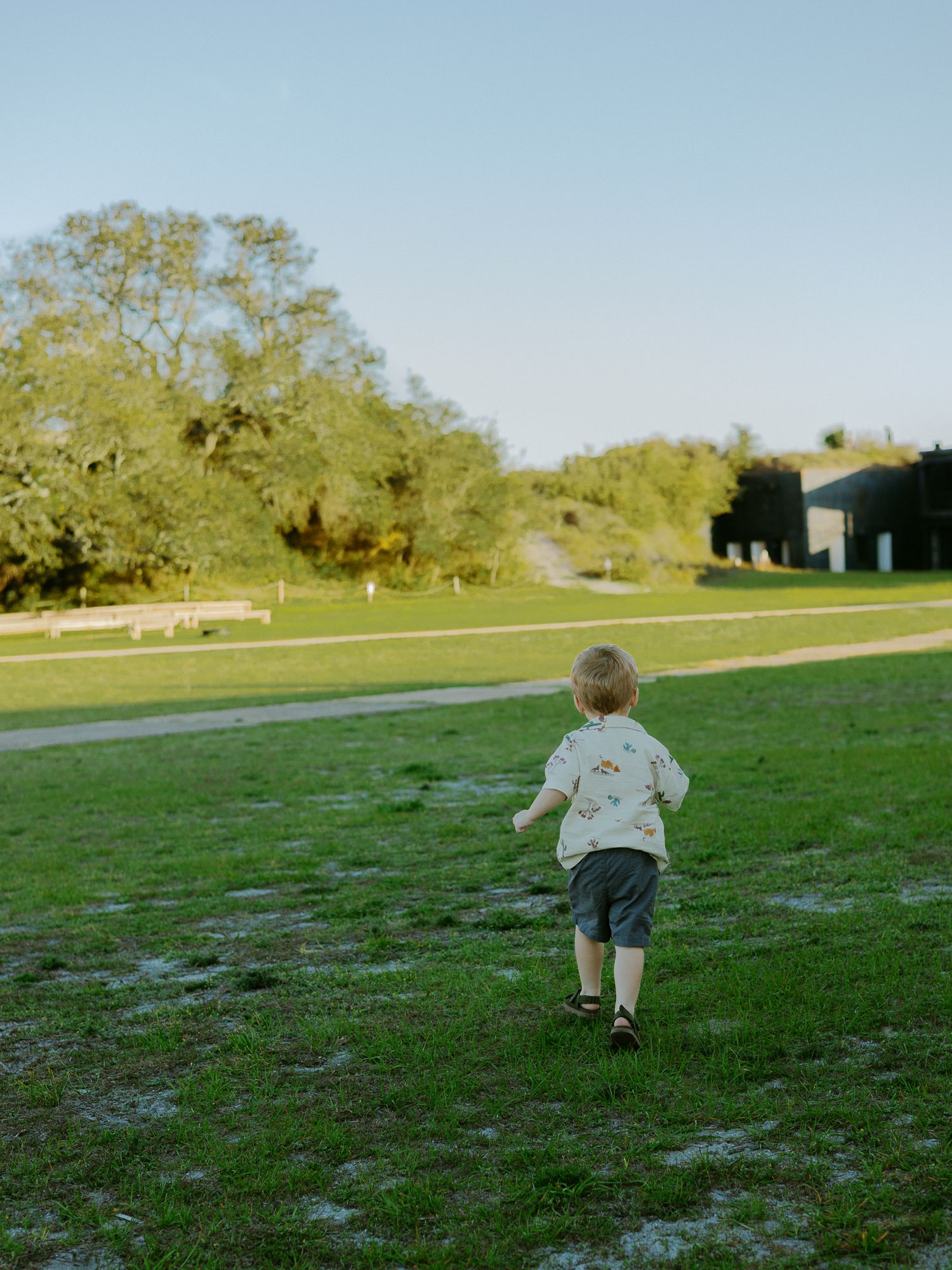 A young toddler runs across a lush green lawn toward trees and a building in warm golden hour light.
