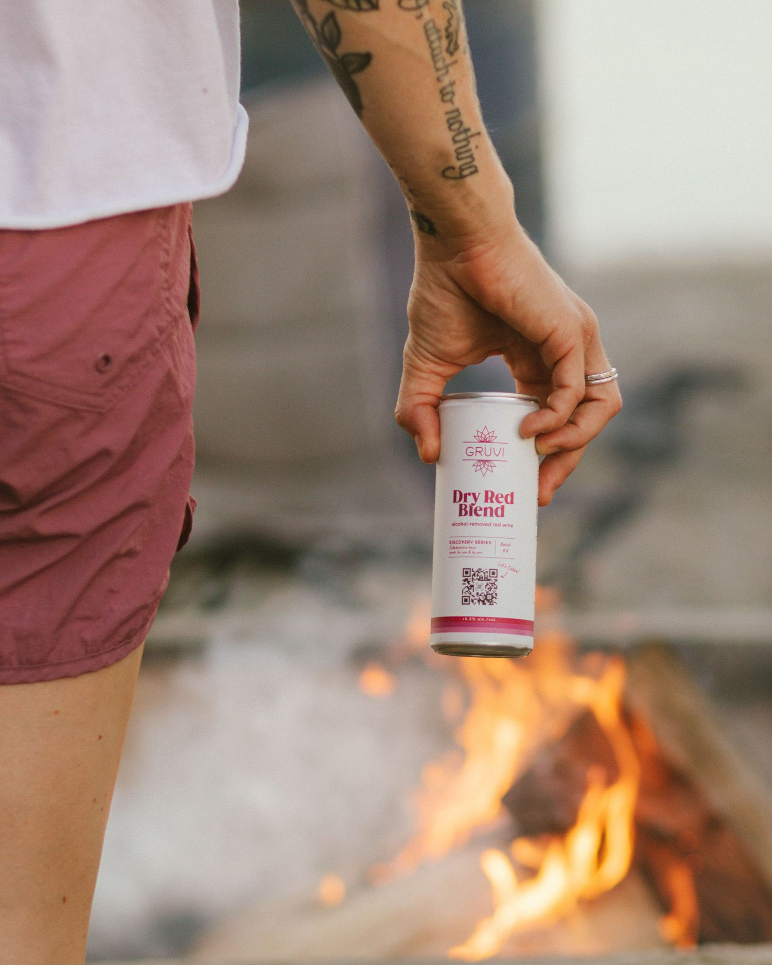 Hand with tattoos holds a small white can near a campfire with flames burning brightly in the background.