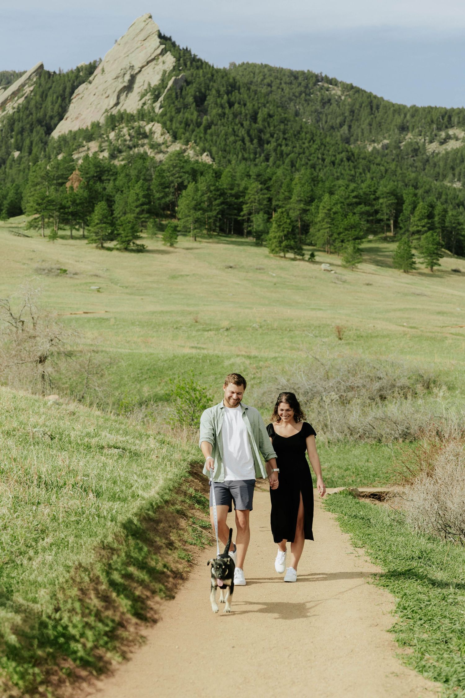 Two people walking a small dog on a hiking trail with the iconic Flatirons rock formation in Boulder, Colorado in the background.
