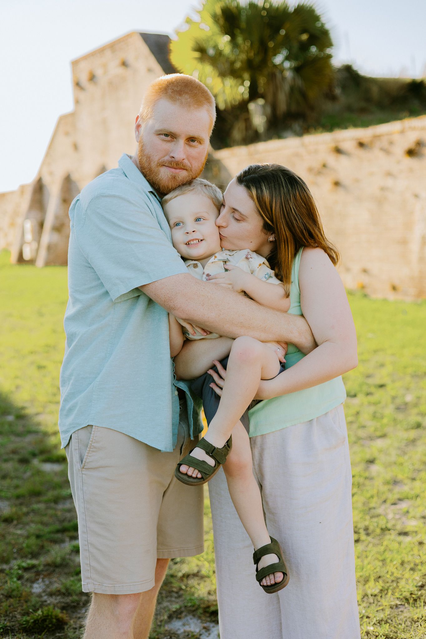 A family of three poses outdoors, father holding toddler while mother kisses child, historic stone ruins in background.