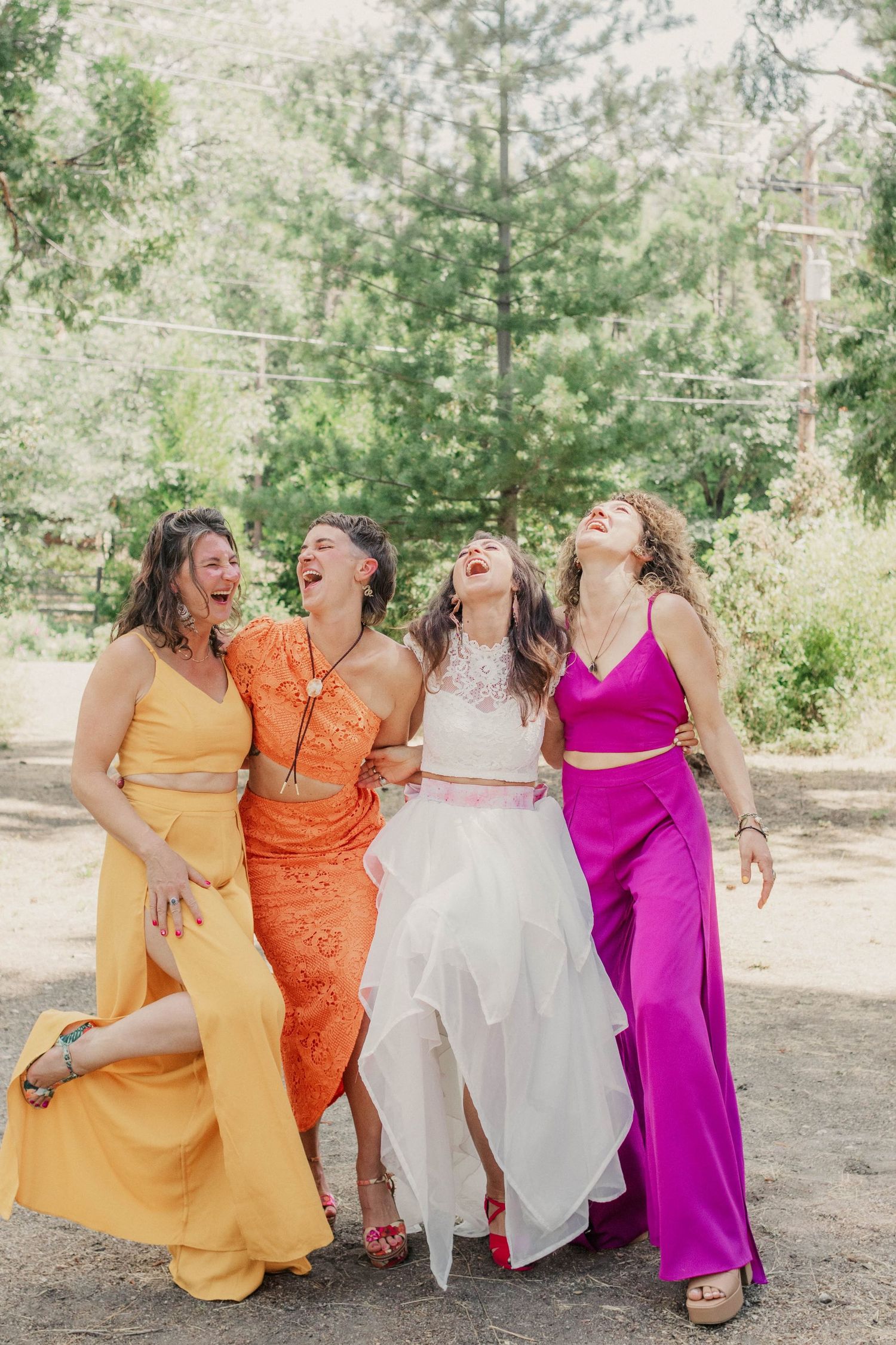 Bride in white dress laughing with three bridesmaids in colorful orange, yellow, and purple outfits outdoors.
