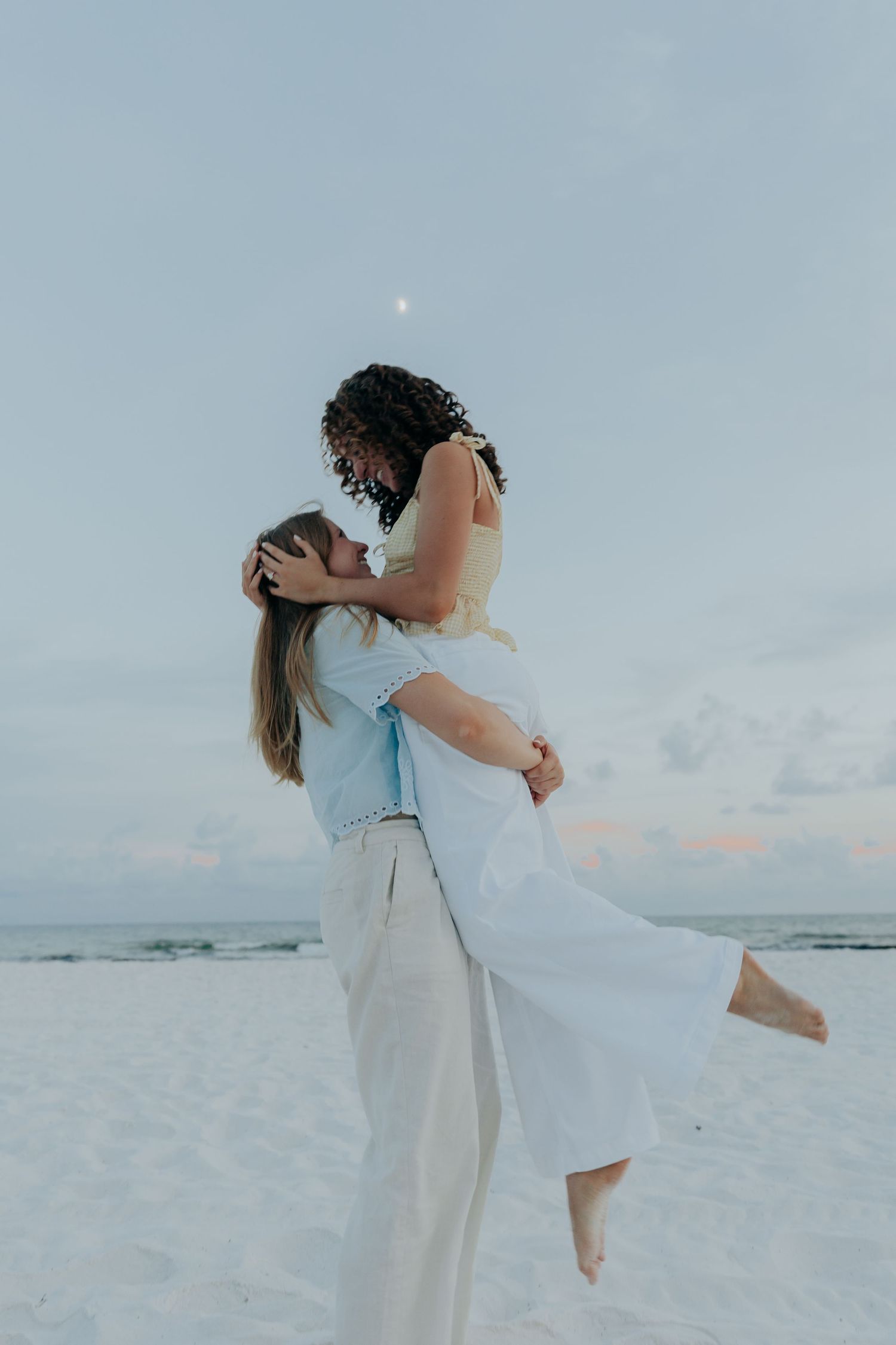 A couple in light beachwear embraces on a white sandy beach during sunset, creating a romantic seaside moment.