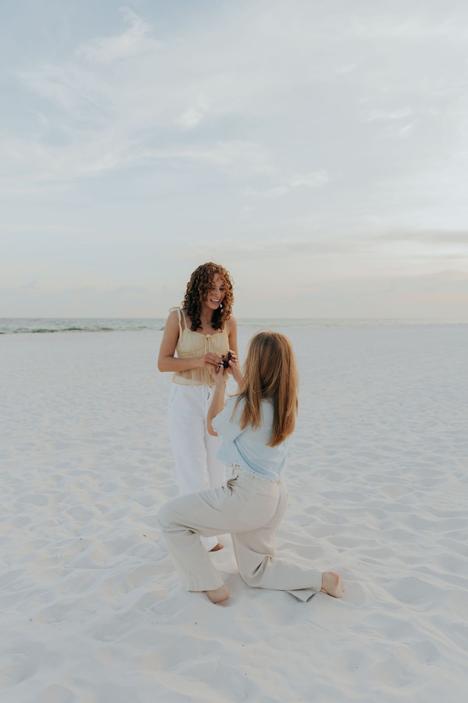 Two people in white outfits embrace on pristine white sand beach during a serene sunset photoshoot.