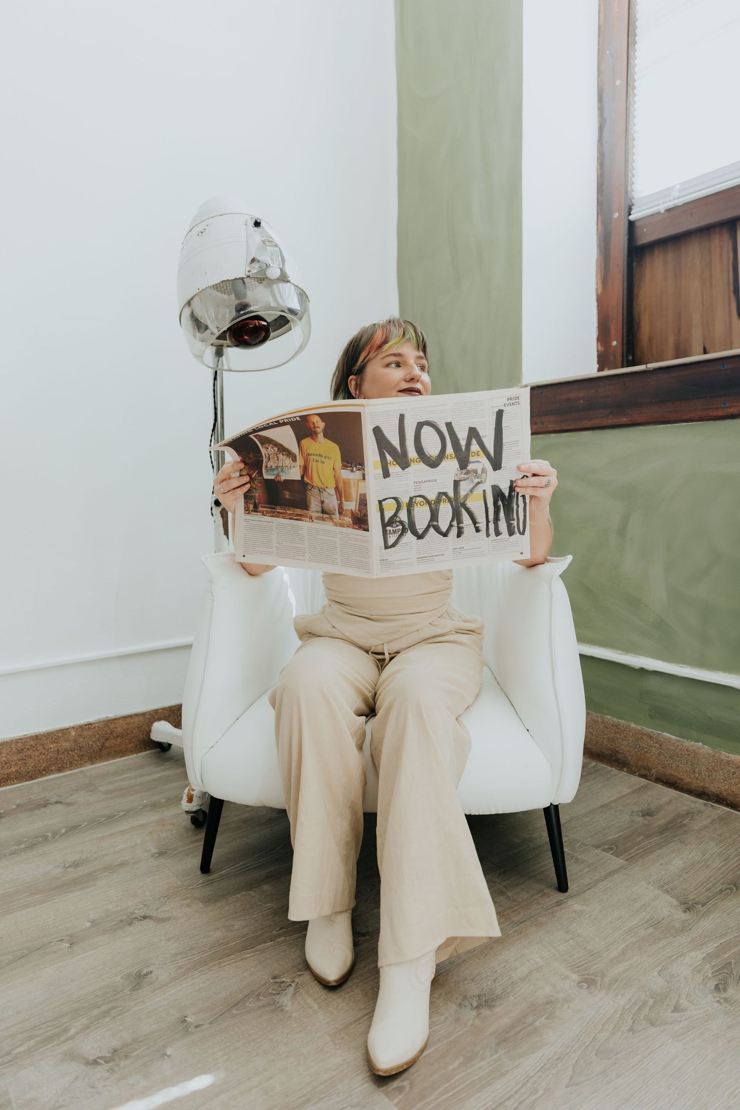Person in white boots and beige pants sits in white chair holding signs that spell out 'now booking' in a minimalist setting.