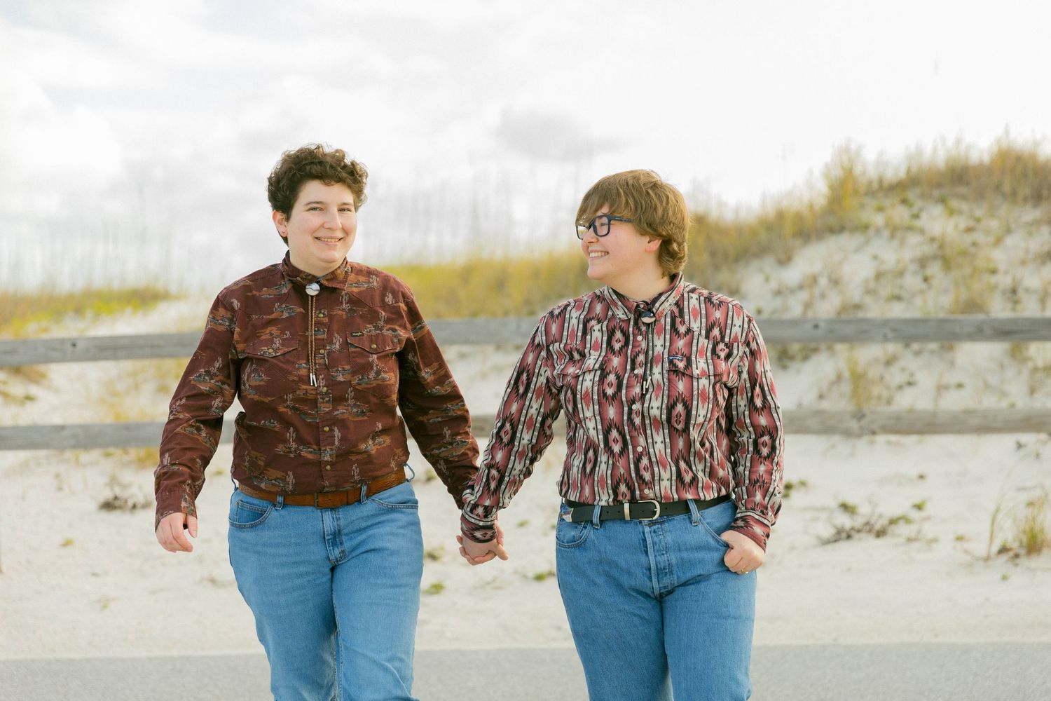 Two people walking hand in hand along a sandy beach path on a cloudy day.