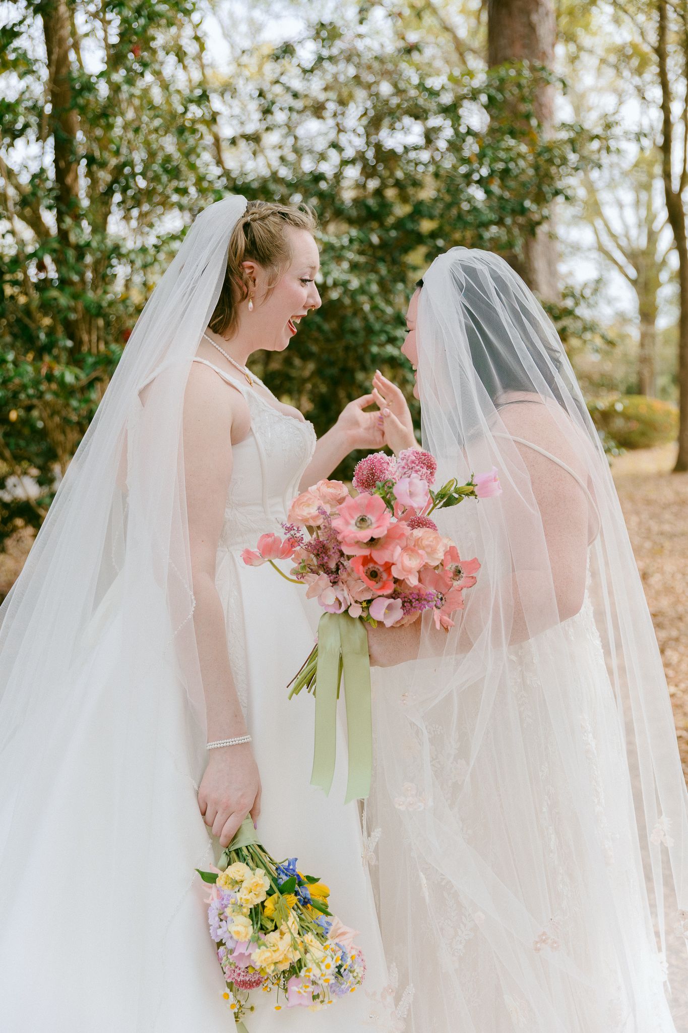 Two brides in white gowns and veils share a tender moment, holding colorful pink and yellow bouquets outdoors.