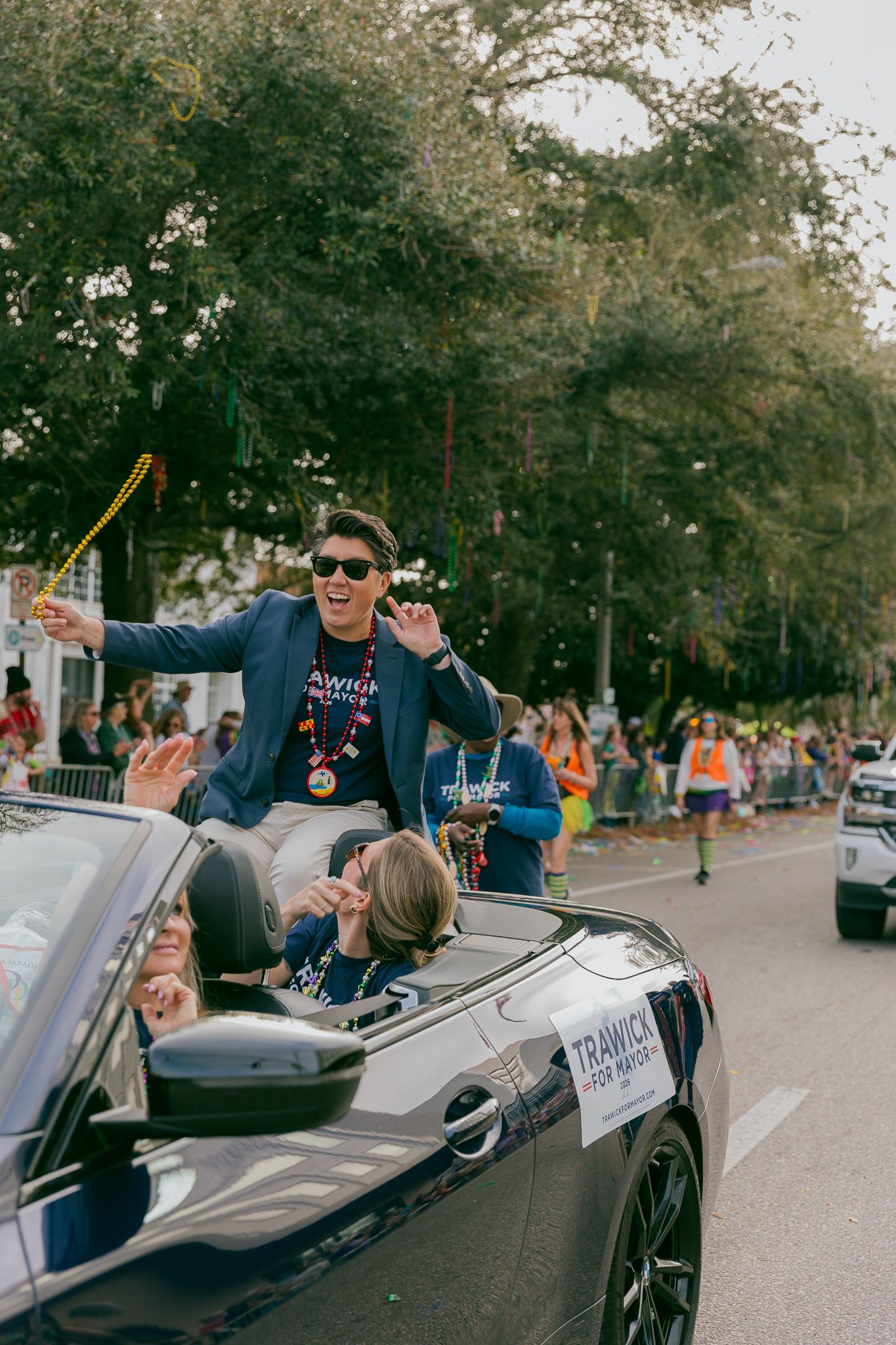 Person waving from a silver BMW convertible during a street parade celebration.