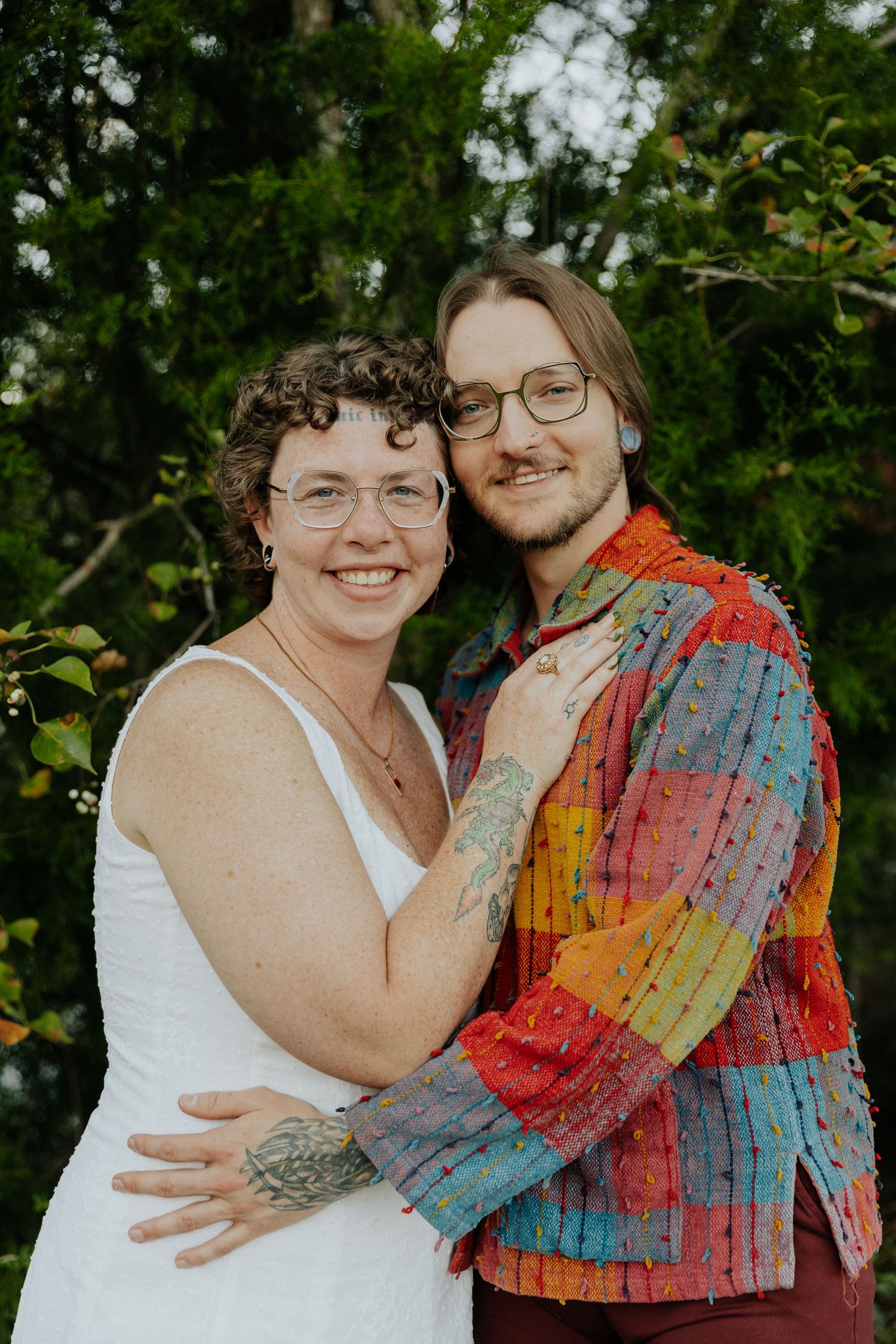 A couple shares a tender embrace outdoors against a backdrop of lush green trees and foliage.