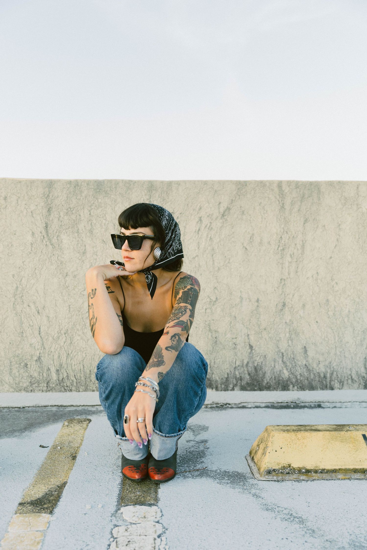 Person in sunglasses and denim sits against a wall in a minimalist urban setting with muted tones.