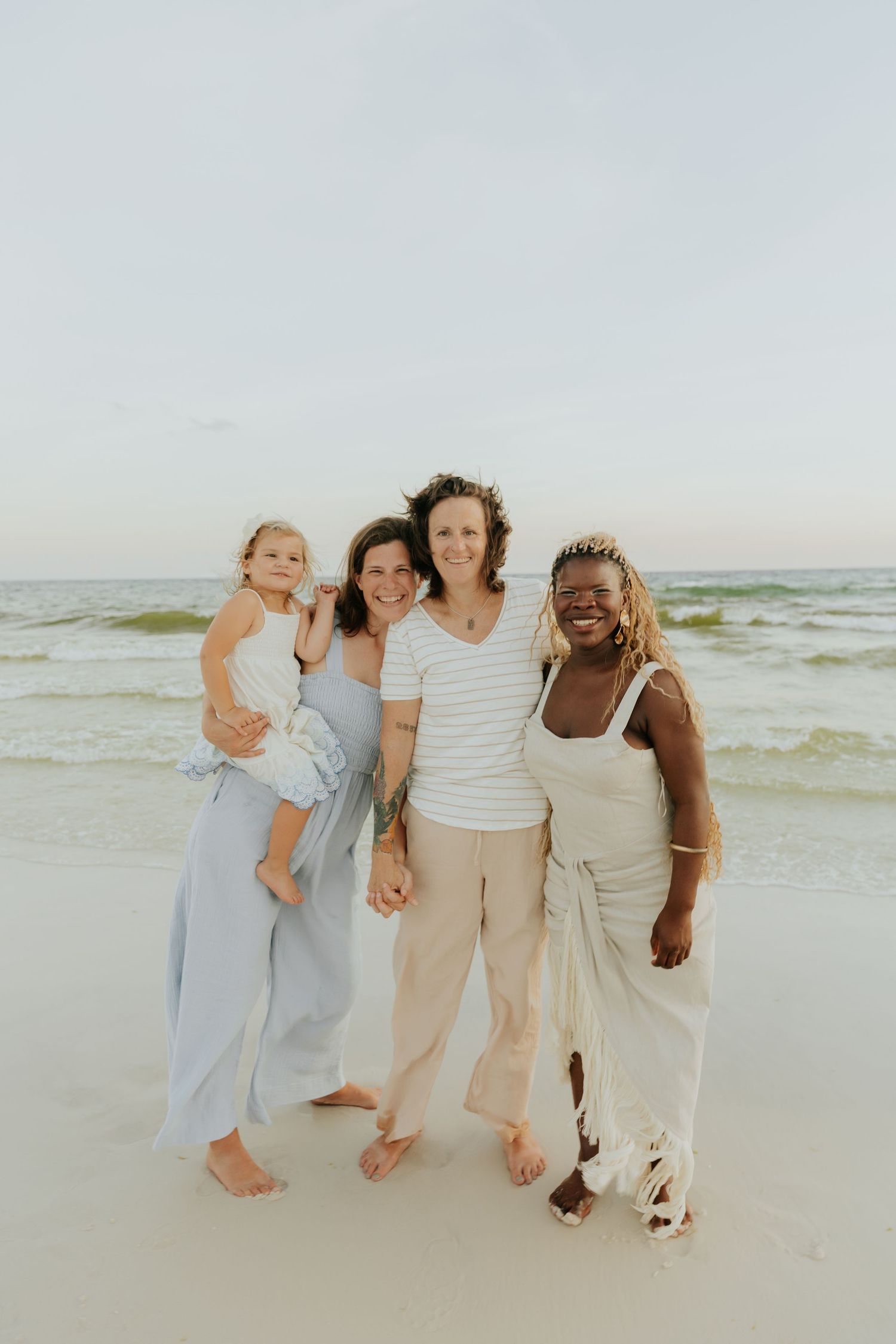 Family in neutral colored clothing embrace while standing together on a sunny beach.