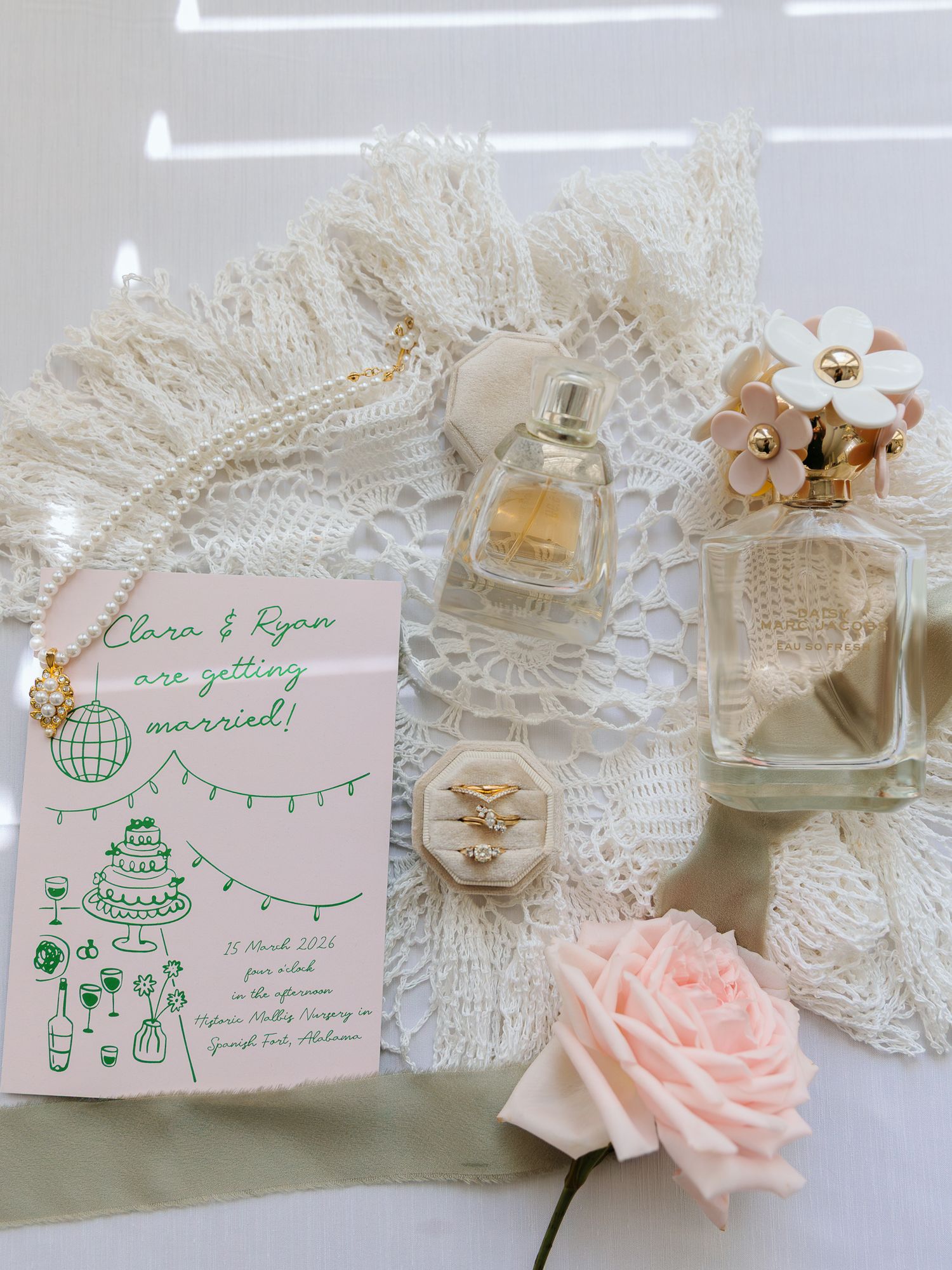 Wedding flat lay with pink invitation, pearl necklace, perfume bottles, ring box, and rose on lace fabric.