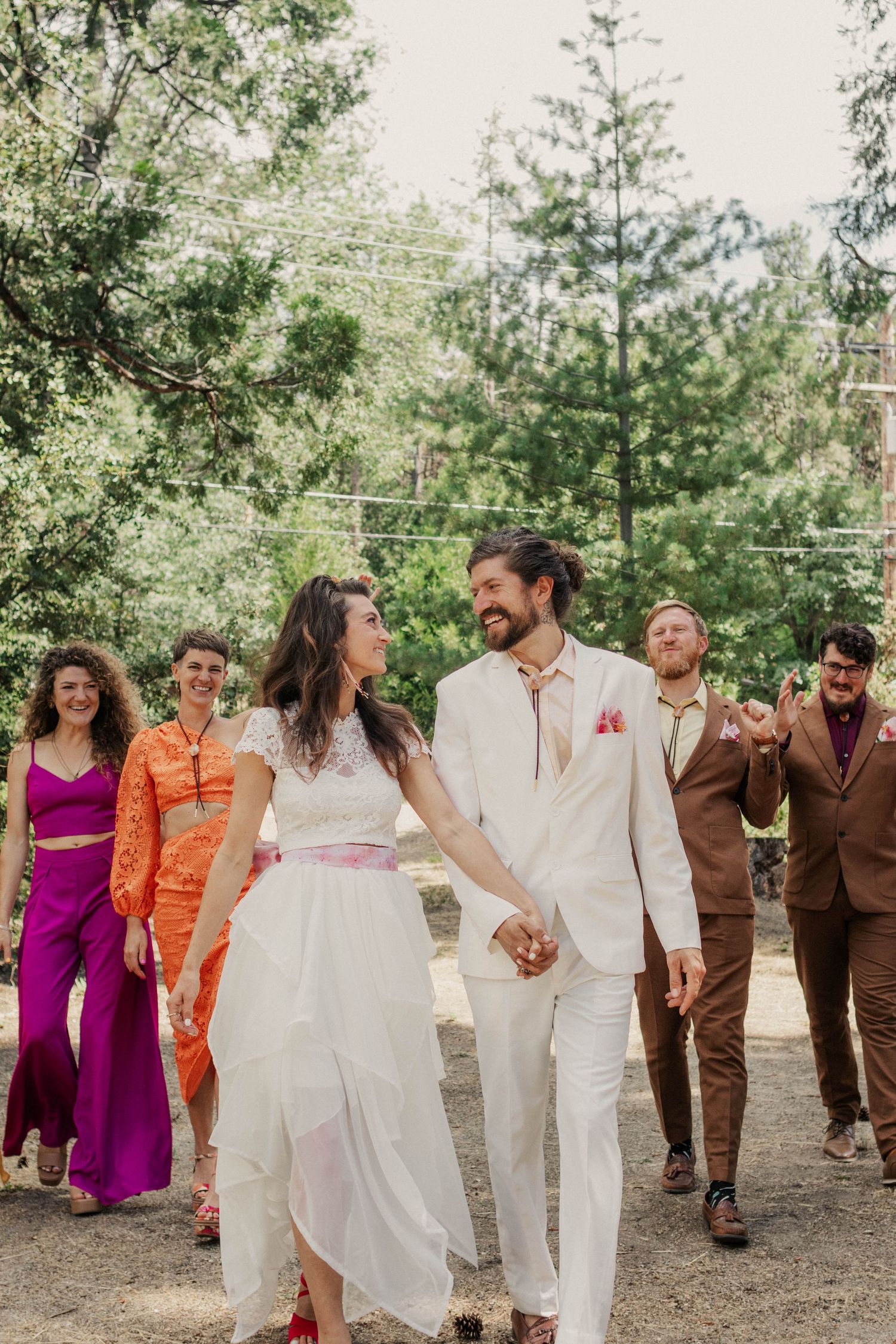 Wedding party walks together on outdoor path with colorful retro-inspired attire and natural forest backdrop.