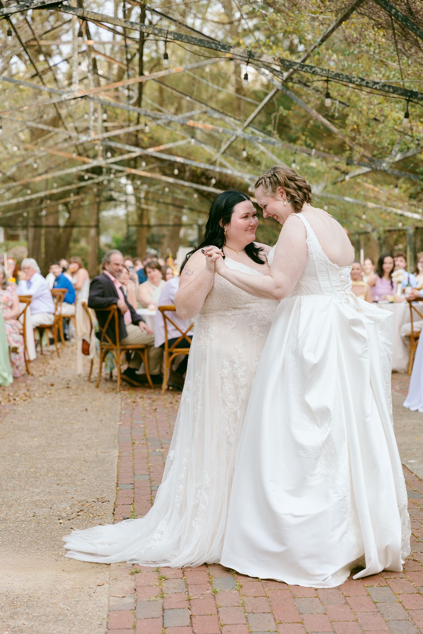 Two brides in white gowns share an intimate first dance at an outdoor wedding reception under string lights.