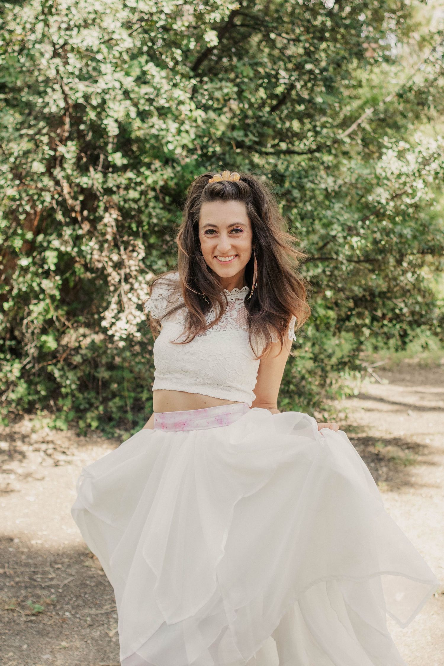 Smiling bride in a two-piece white lace wedding dress twirling outdoors among lush green trees.
