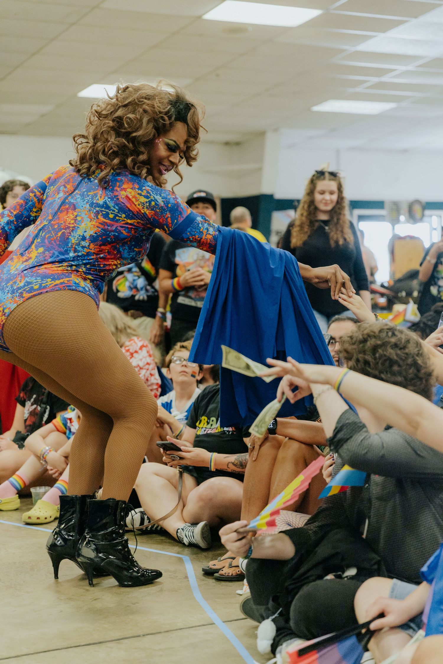 Performer in colorful leotard entertaining crowd during indoor show with blue fabric prop.