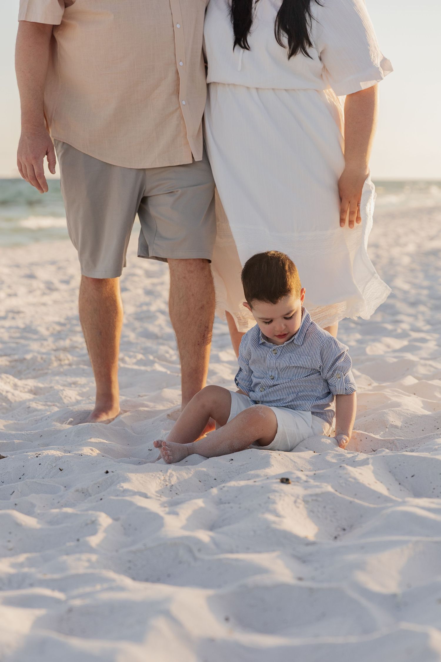 A child plays in soft white sand at the beach while adults stand nearby in light-colored casual clothing.