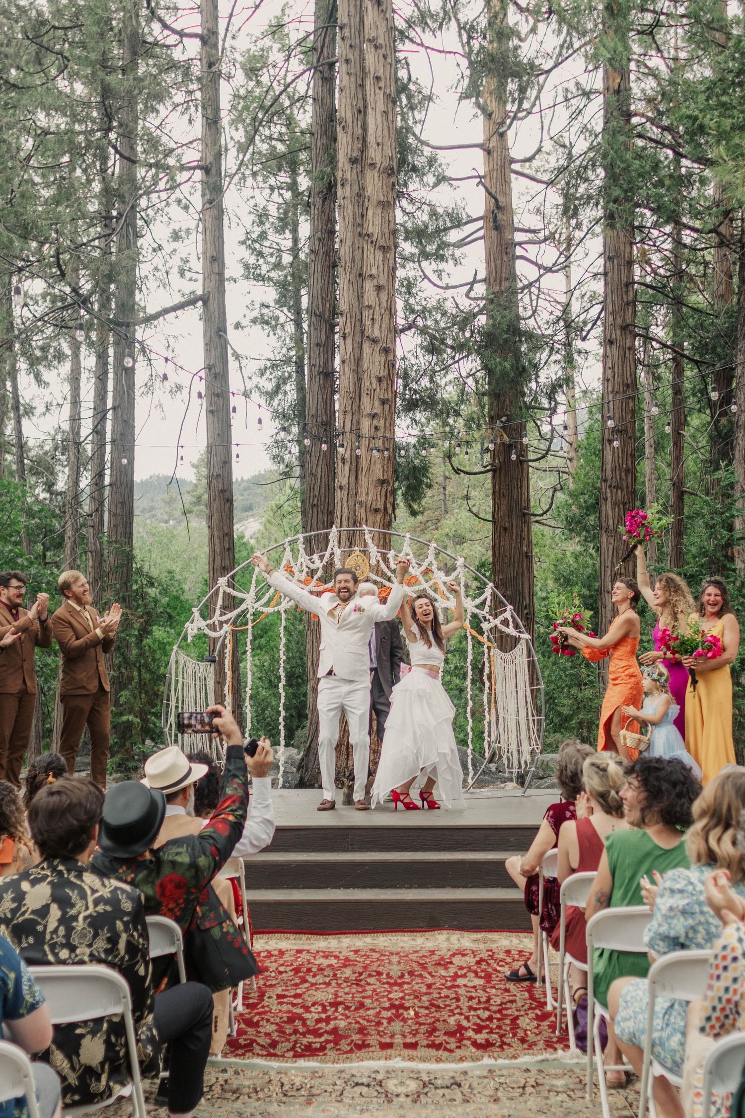 Joyful couple celebrates wedding ceremony exit under macrame arch surrounded by towering redwoods and cheering guests.