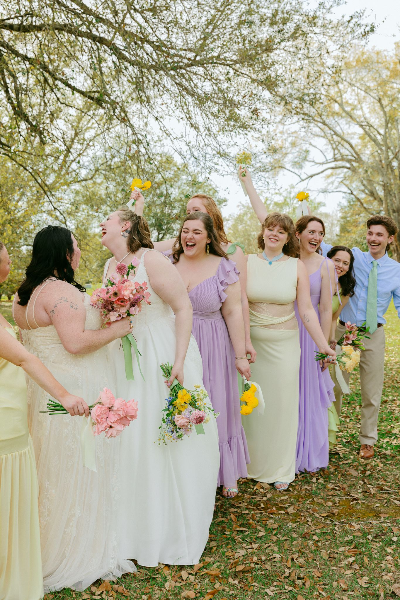 Bridesmaids in pastel purple and yellow gowns laugh and celebrate with bride outdoors at a wedding.