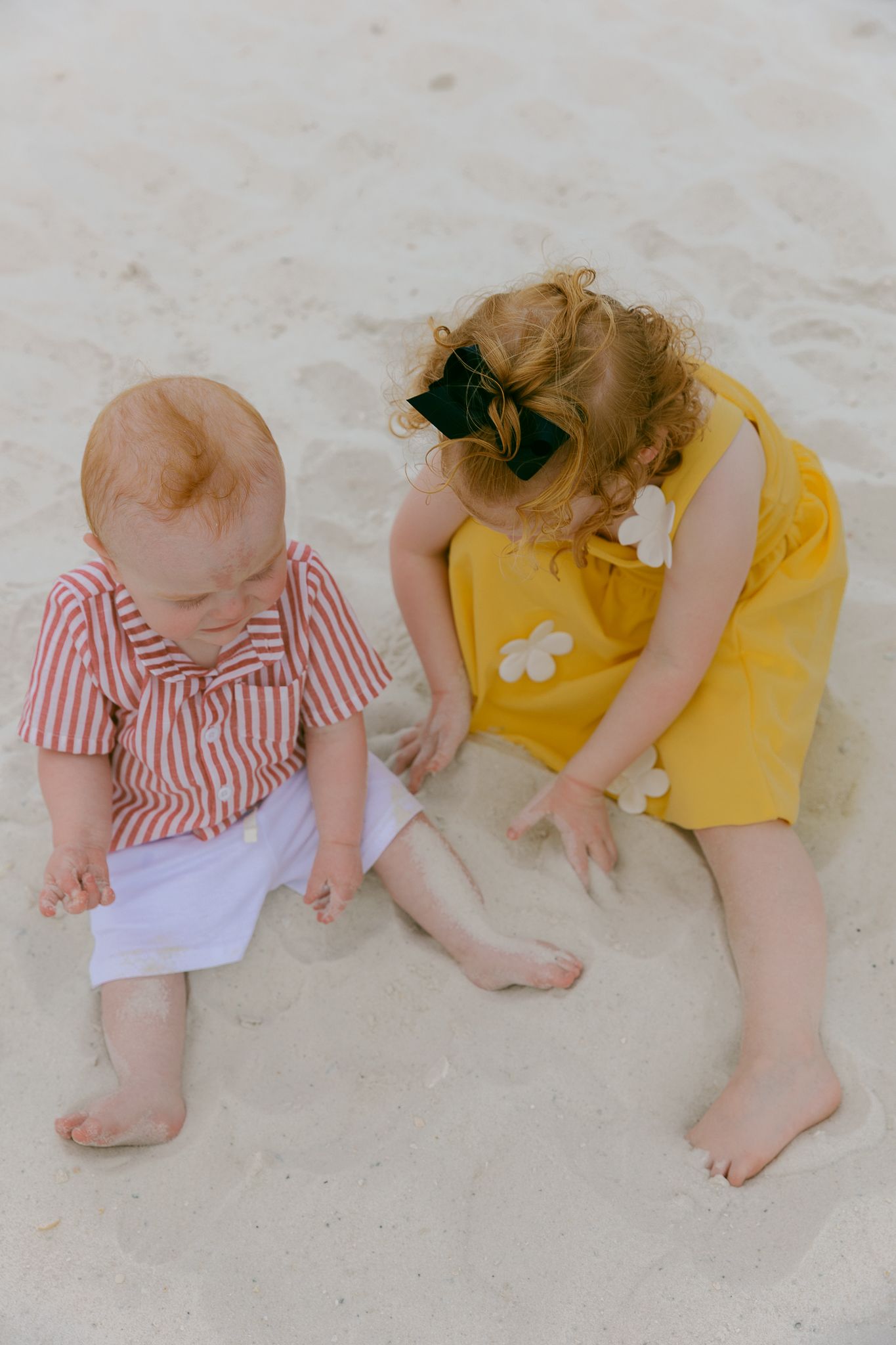 Two babies playing in white sand at the beach, one in red stripes, one in a yellow dress with flowers.