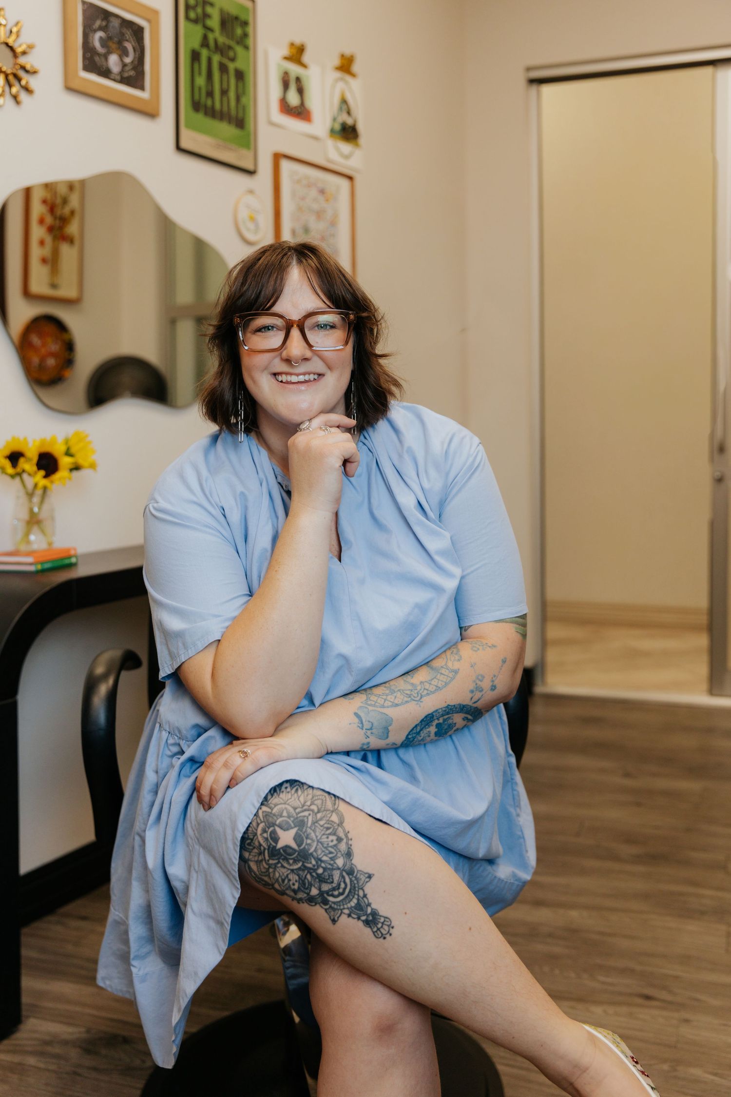 A person in a light blue dress sits casually in a room with artwork and sunflowers, showing off a detailed leg tattoo.