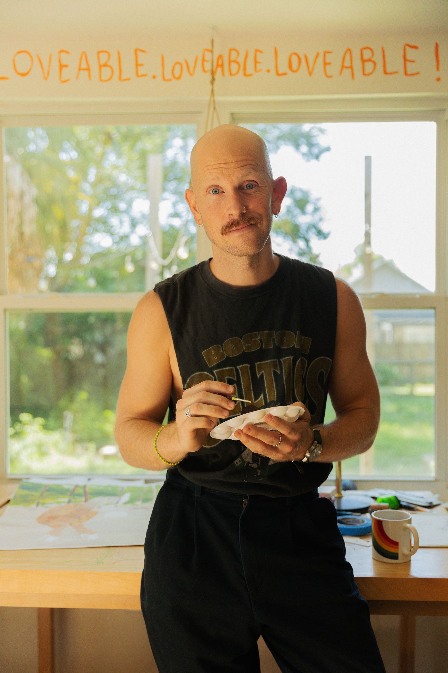 Person in a black sleeveless shirt stands by a window with 'LOVEABLE' text written above in an orange hue.