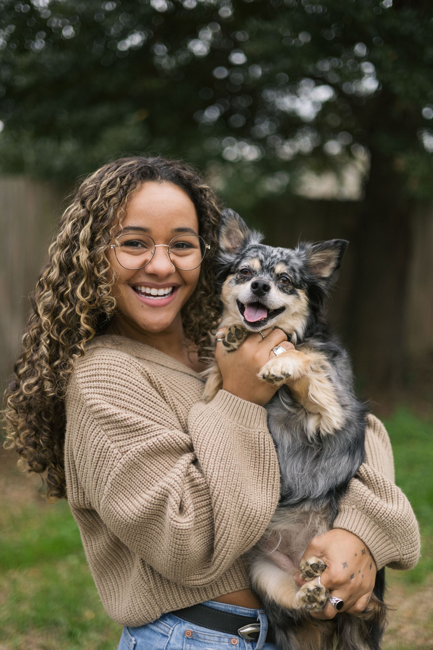 A person in a beige sweater holds and plays with a happy Chihuahua mix dog outdoors in a park setting.